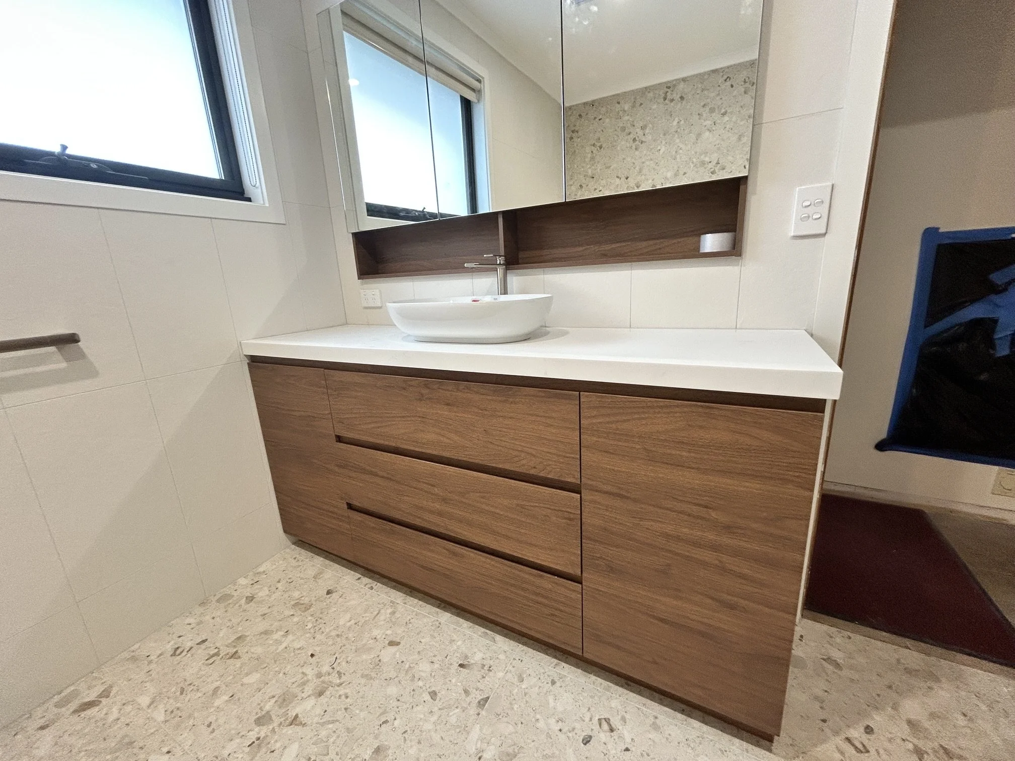 A modern bathroom vanity with a white countertop, a vessel sink, wooden cabinets, and a mirror with a wooden frame. There are windows above the vanity and a power outlet on the wall.