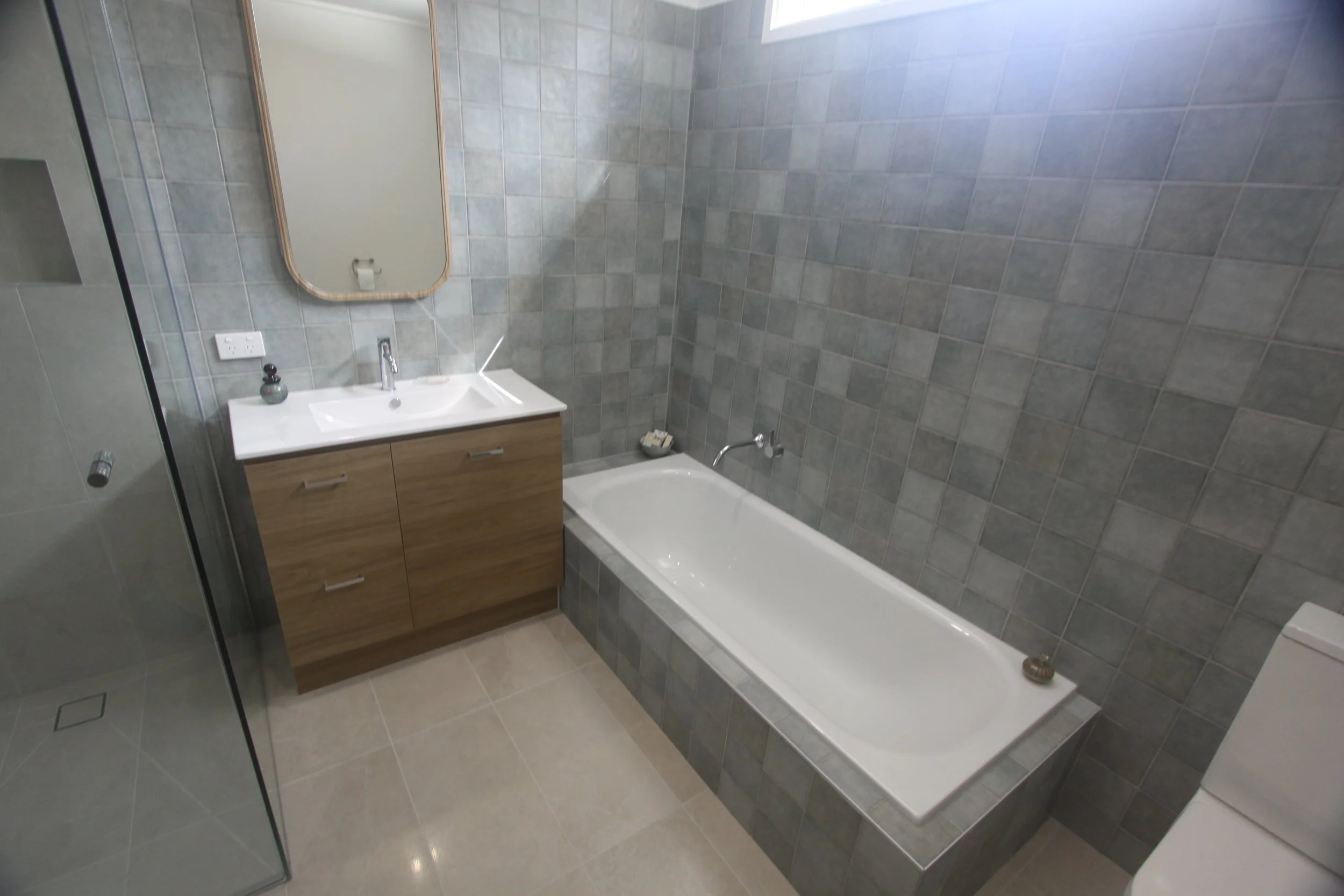 Modern gray-tiled bathroom with a white bathtub, wood vanity, sink, mirror, and glass shower enclosure