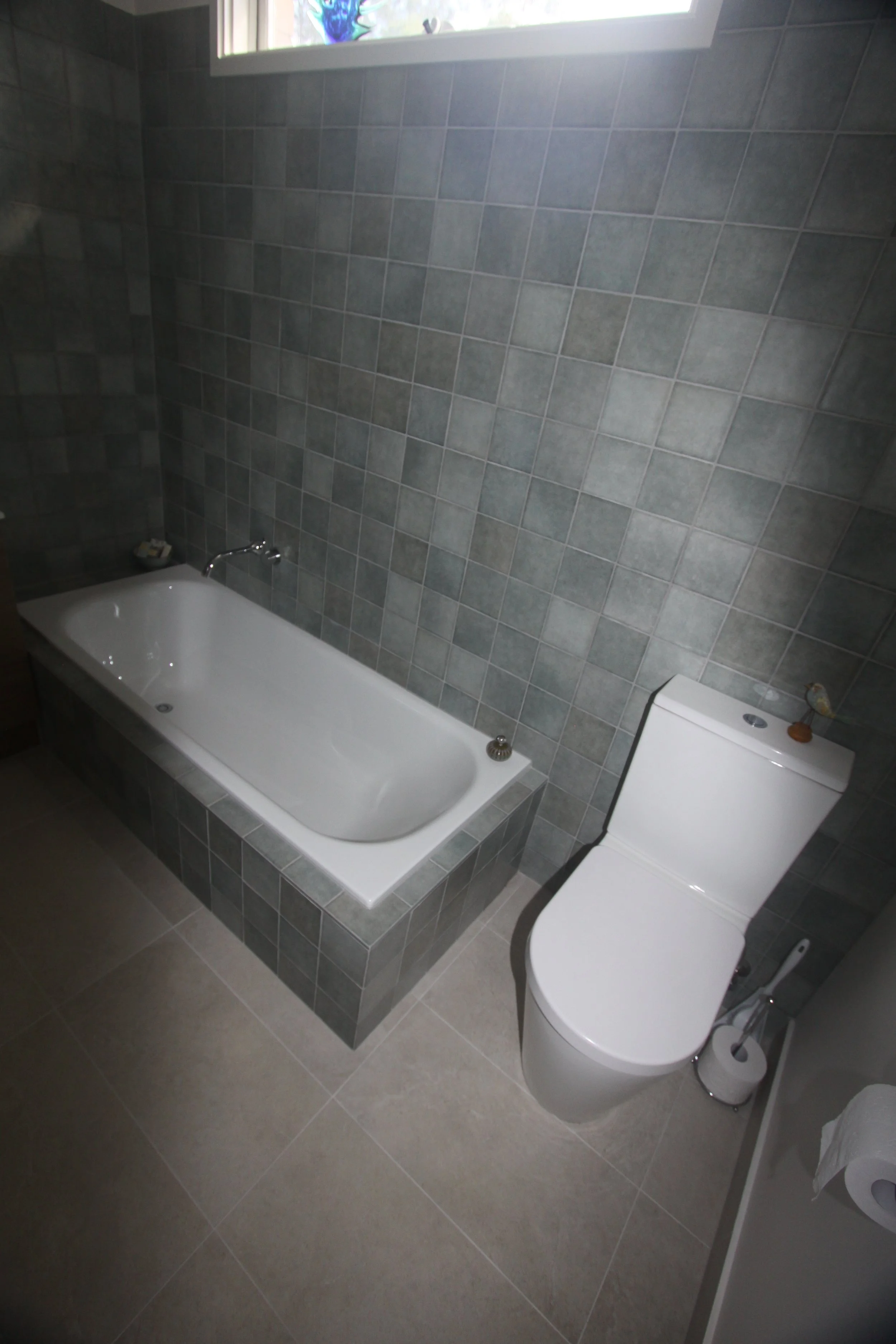 Gray tiled bathroom with a white bathtub, wall-mounted faucet, toilet, and window.