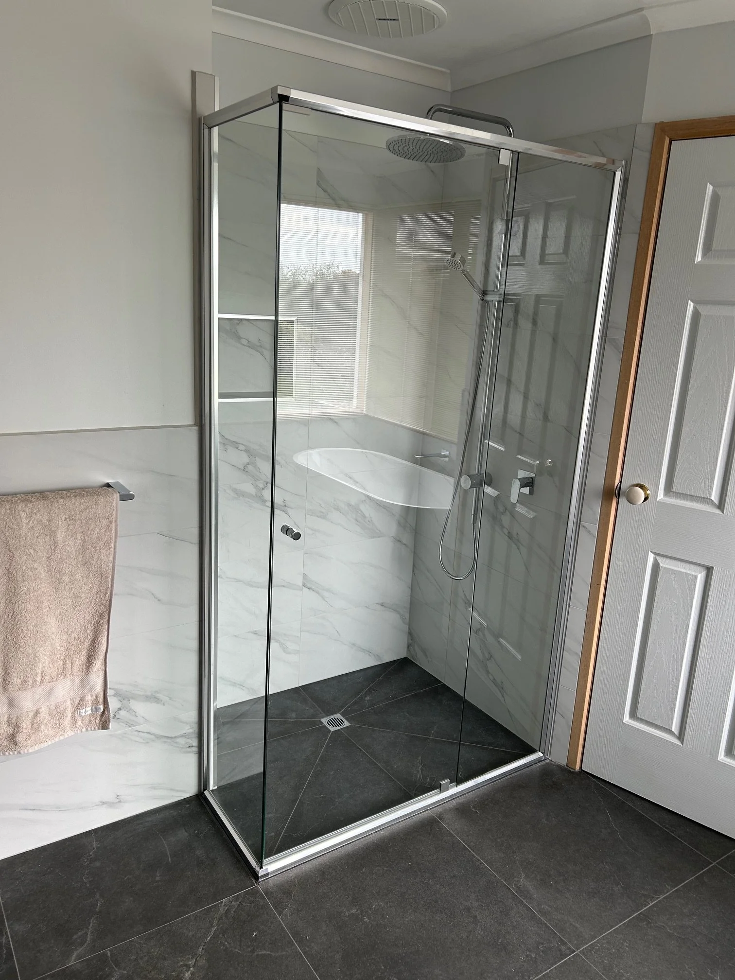 Modern bathroom with a frameless glass walk-in shower, rainfall showerhead, handheld sprayer, white marble-look walls, and dark floor tiles