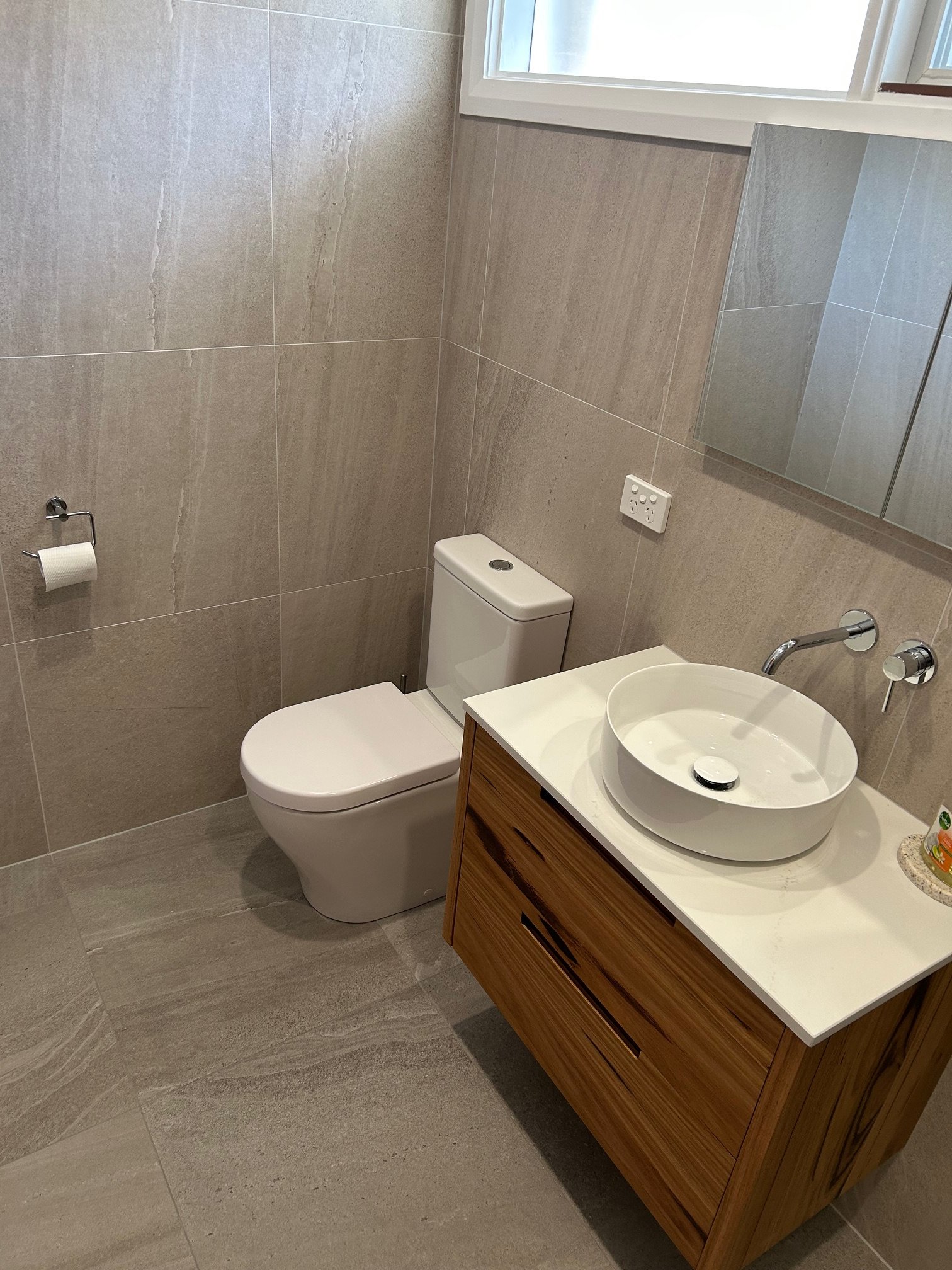 Modern bathroom with a white toilet, round vessel sink, wood vanity, tiled walls, mirror, and window.