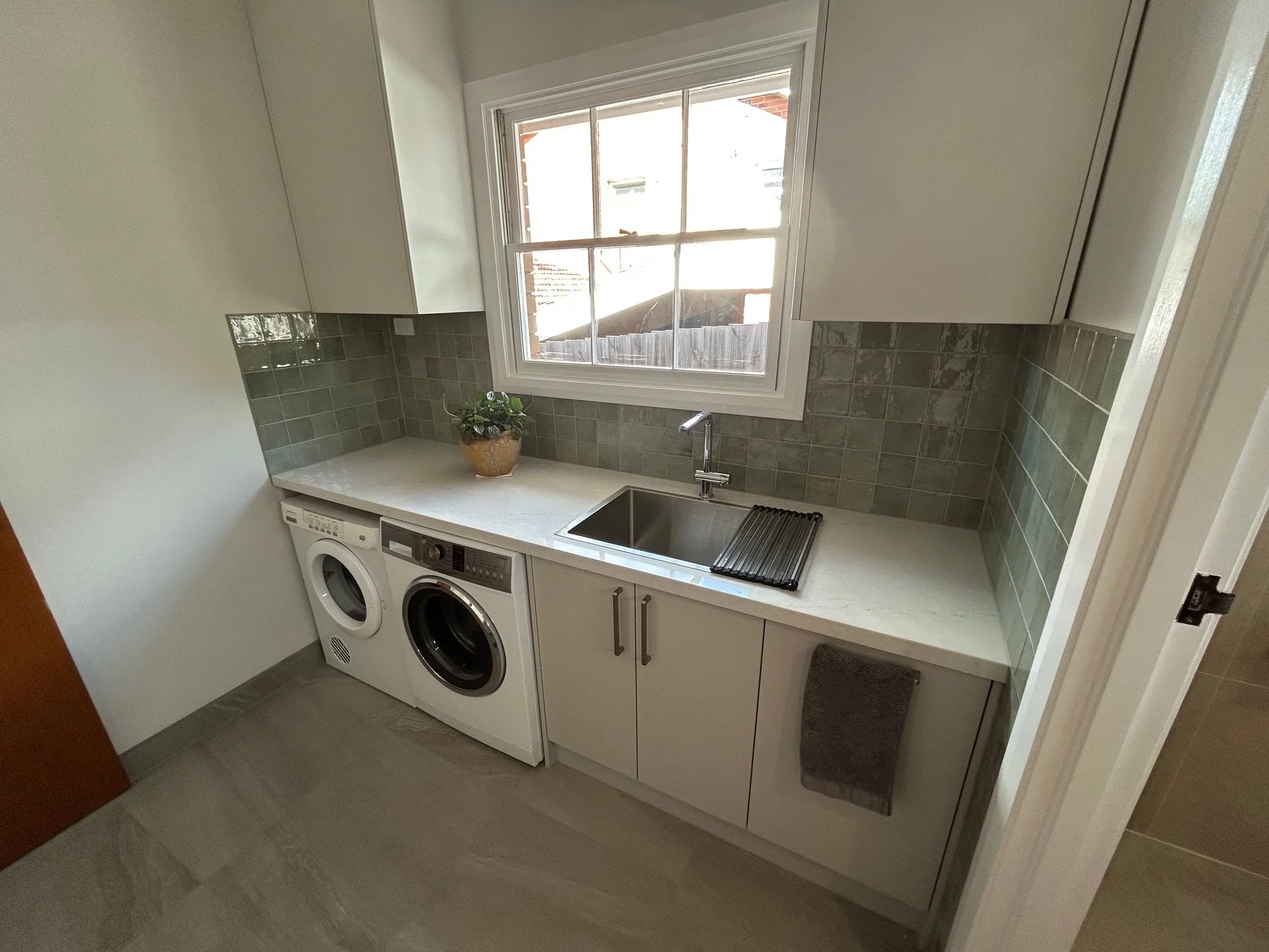 Compact laundry room with washer, dryer, white cabinets, tiled backsplash, countertop sink, drying rack, and window