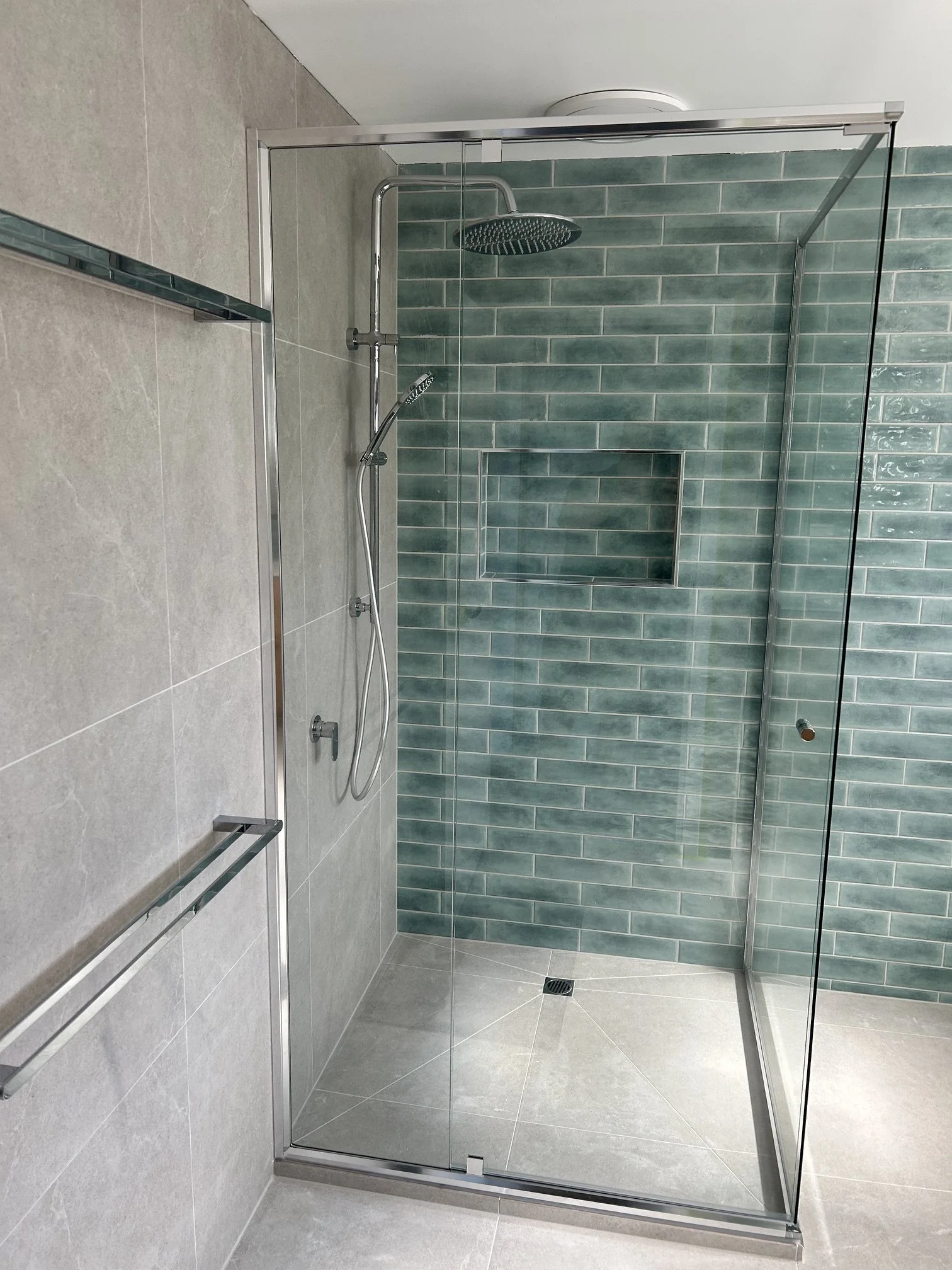 Modern glass-enclosed walk-in shower with green subway tile, rainfall showerhead, handheld sprayer, built-in niche, and gray tile floor