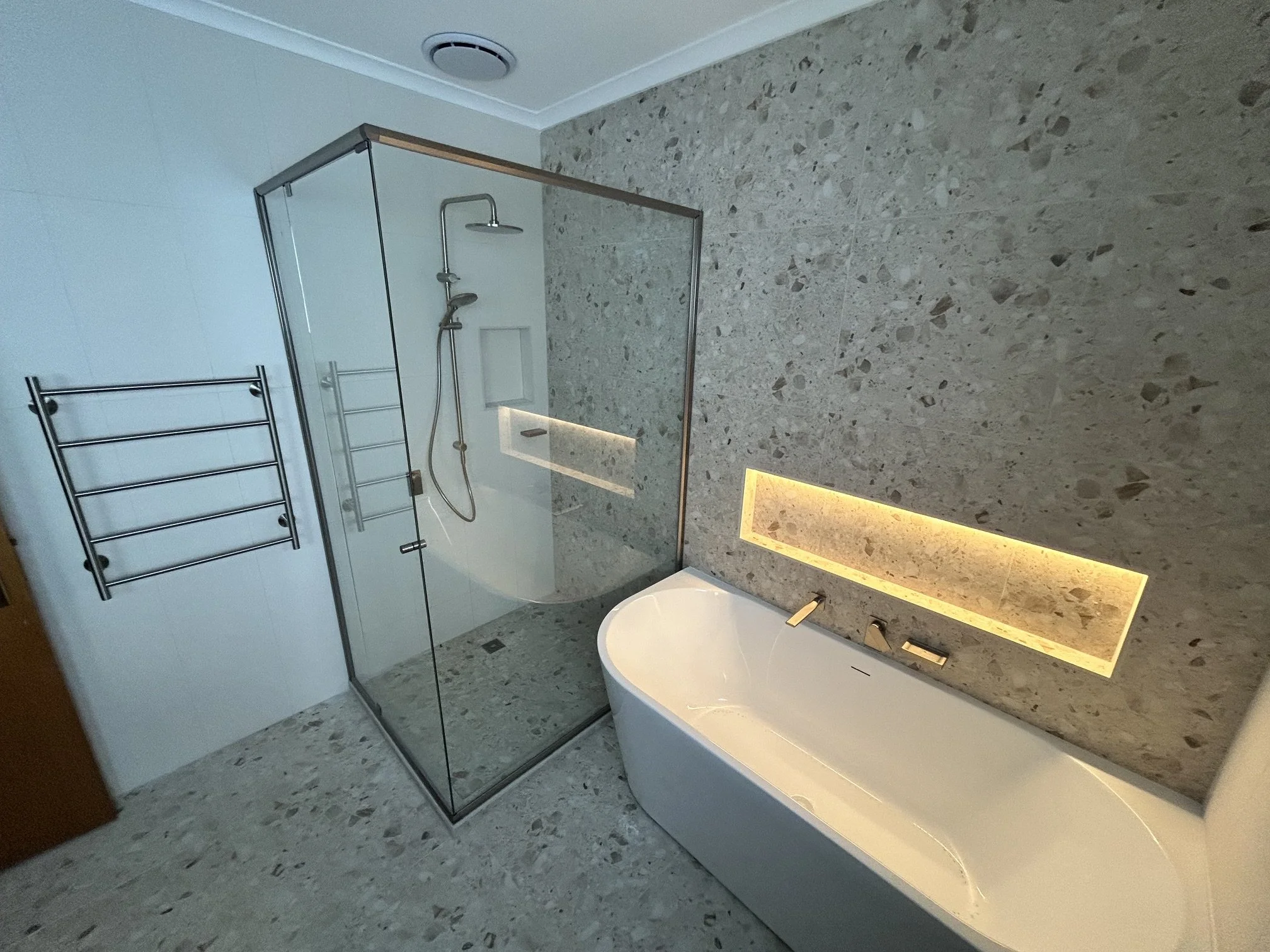 A modern bathroom with a walk-in shower enclosed by glass, a rain showerhead, a hand shower, a wall niche with lighting, a white bathtub, and a towel rack, all with speckled gray tiles.