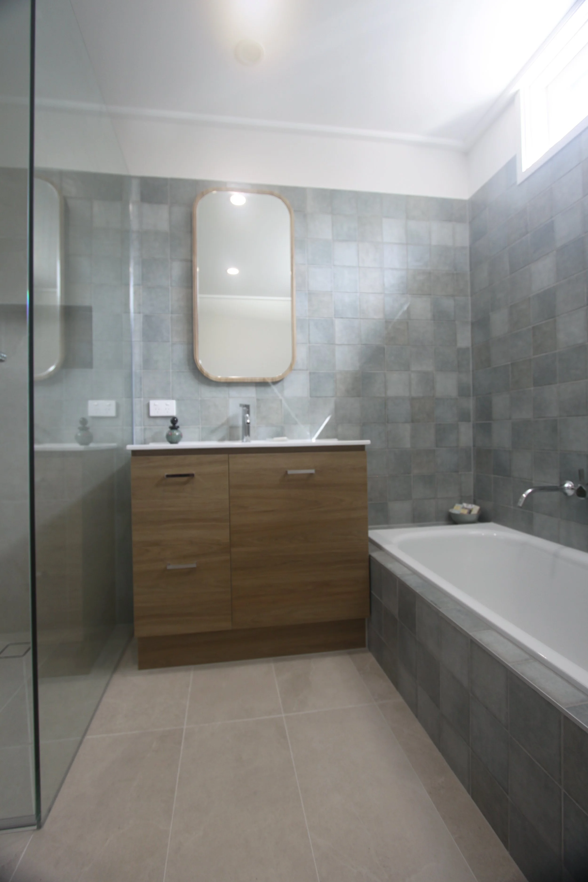 Modern bathroom with gray tiled walls, wood vanity, rounded rectangular mirror, bathtub, and glass shower enclosure