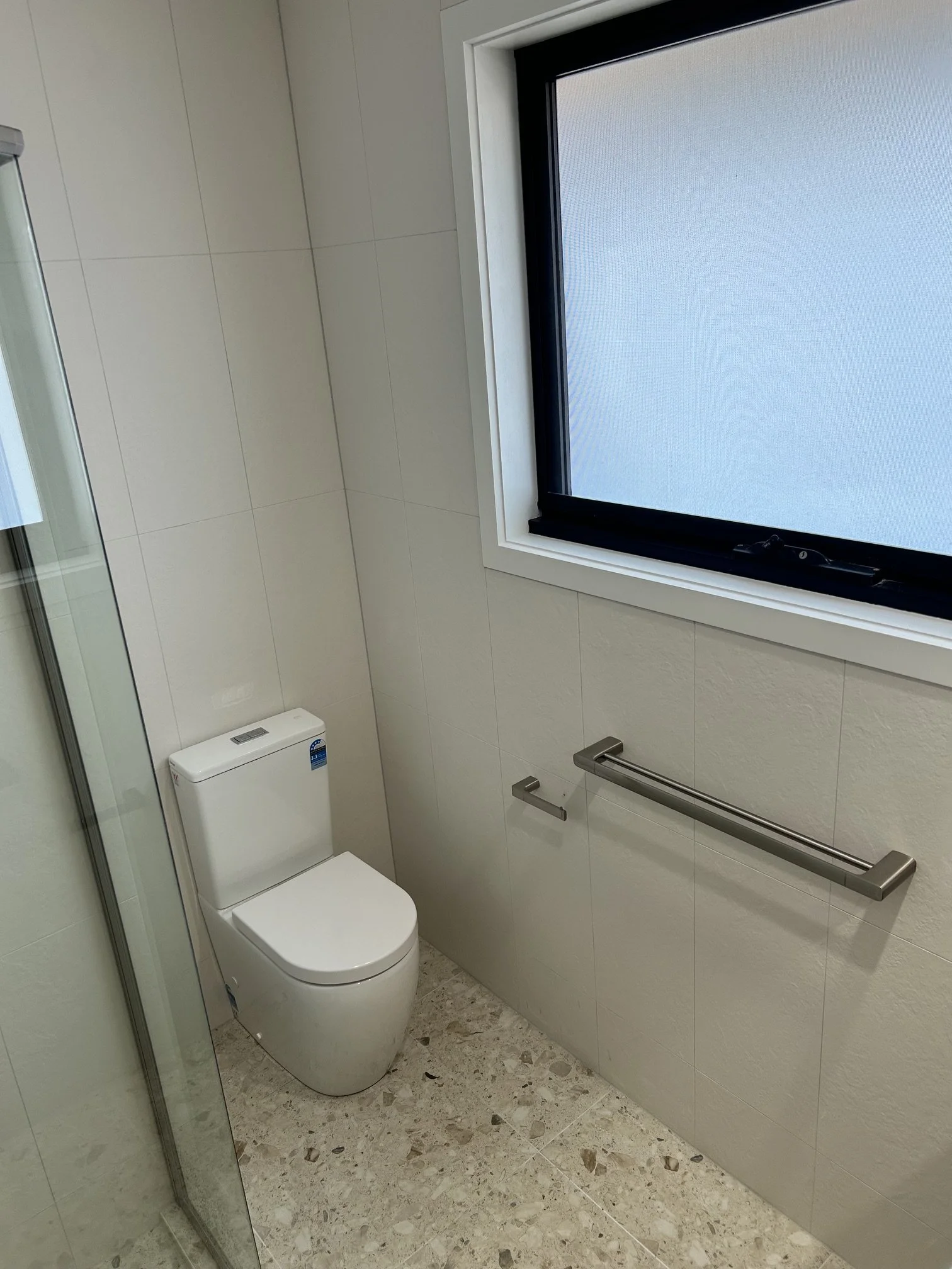 A modern bathroom with a white toilet, beige tiled floor, and a large frosted window with a black frame. There are wall-mounted grab bars near the toilet.