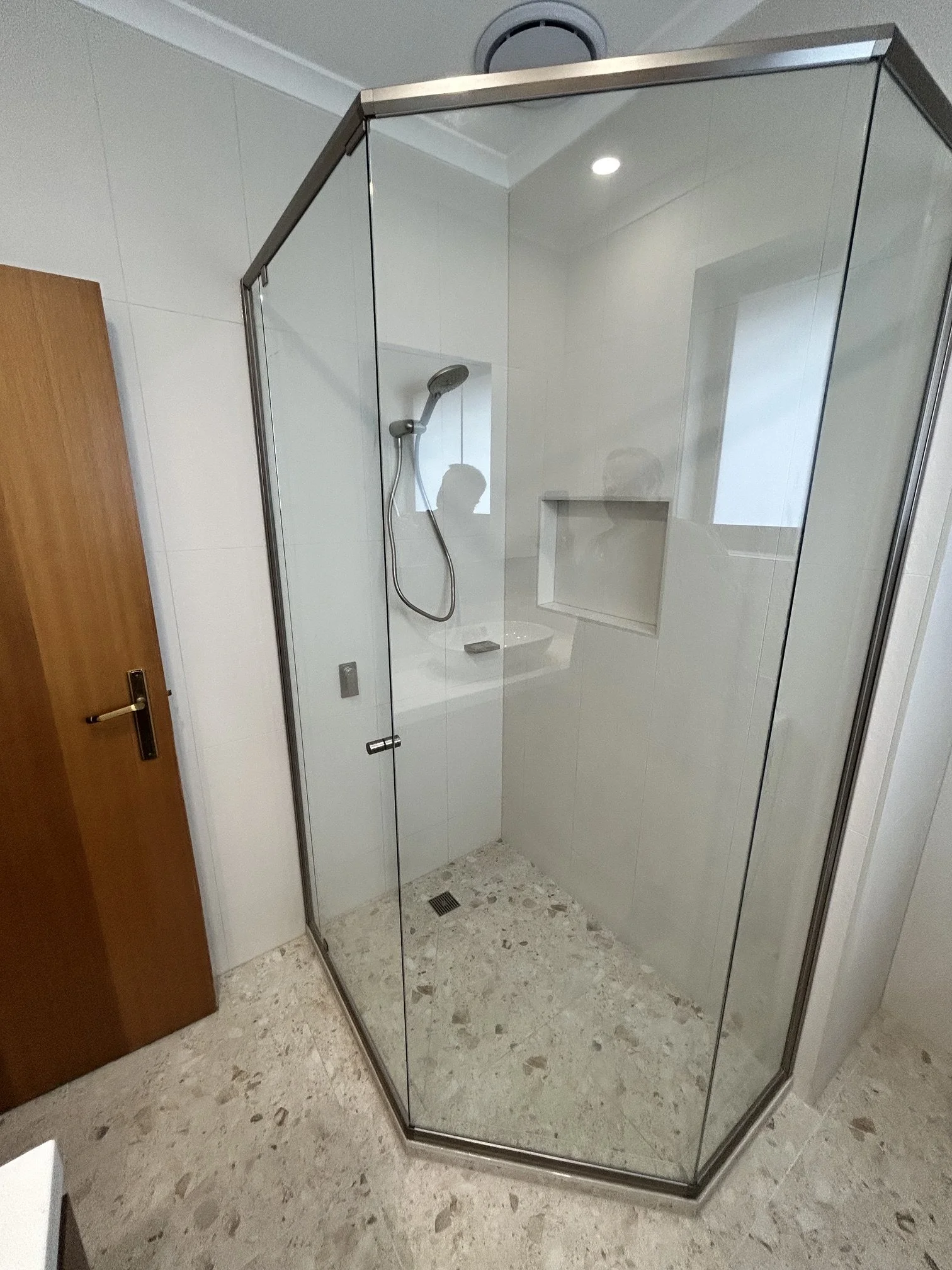 A modern corner shower with glass walls, a handheld showerhead, a built-in niche, tiled walls, and marble flooring in a bathroom.