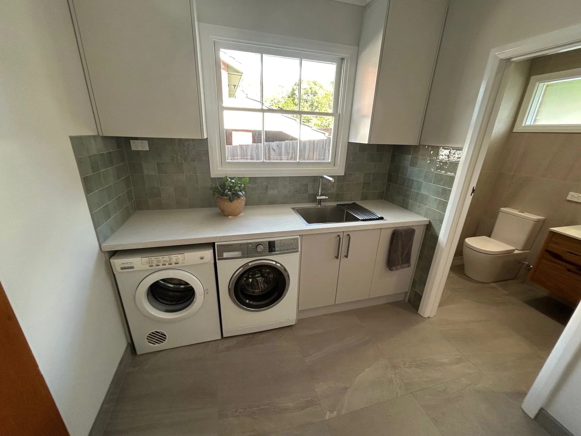 Modern laundry room with washer, dryer, sink, tiled backsplash, storage cabinets.