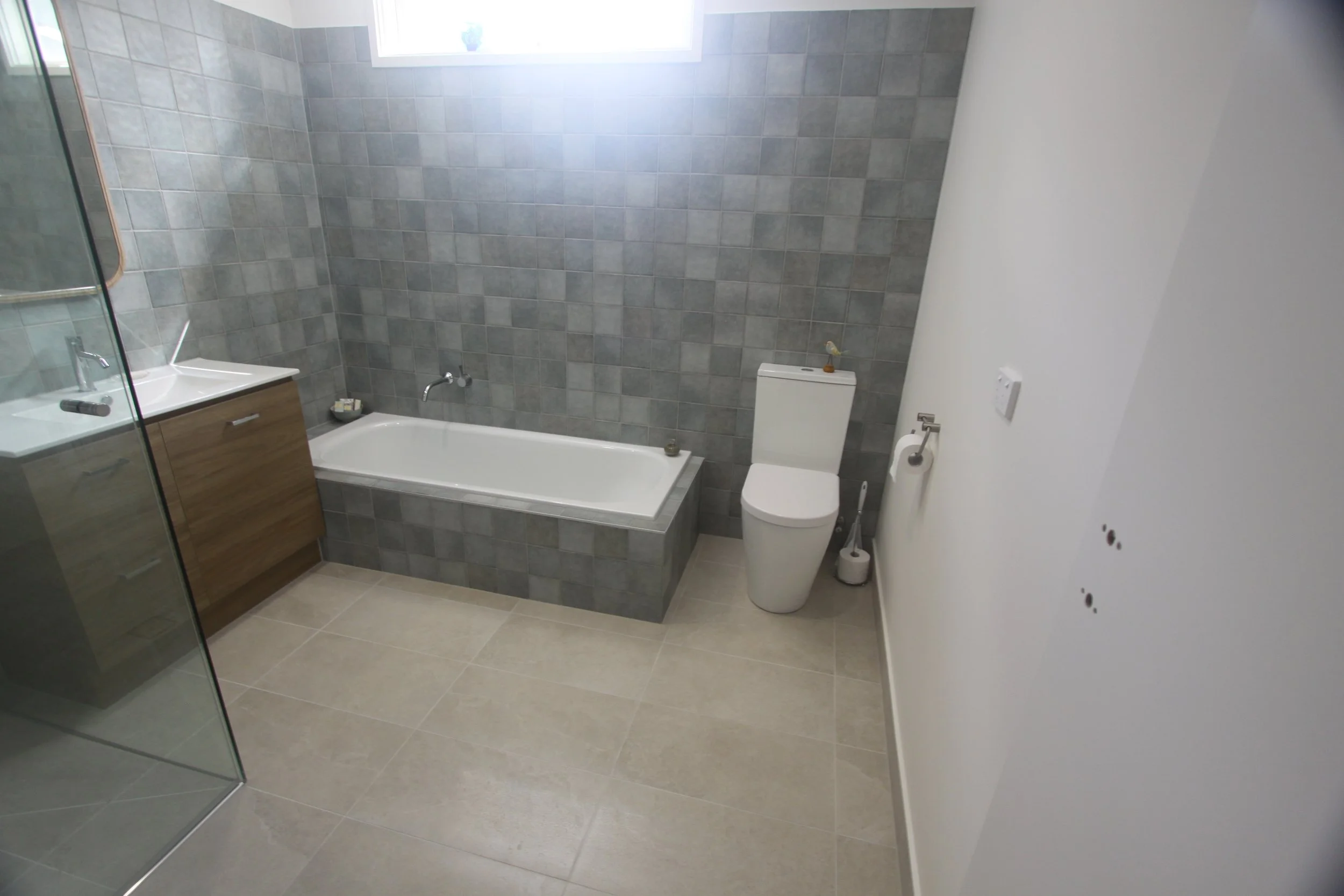 Modern bathroom with gray tiled walls, bathtub, glass shower enclosure, wood vanity, sink, and toilet.