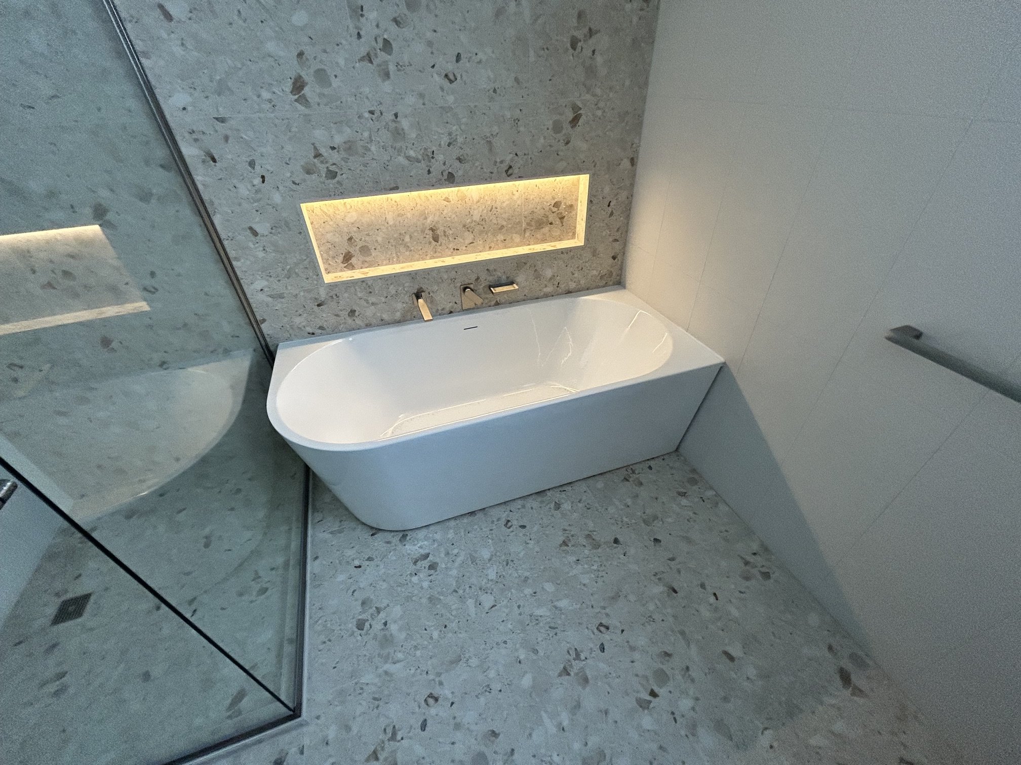 Modern white freestanding bathtub in a terrazzo-tiled bathroom with glass shower enclosure and backlit recessed wall shelf
