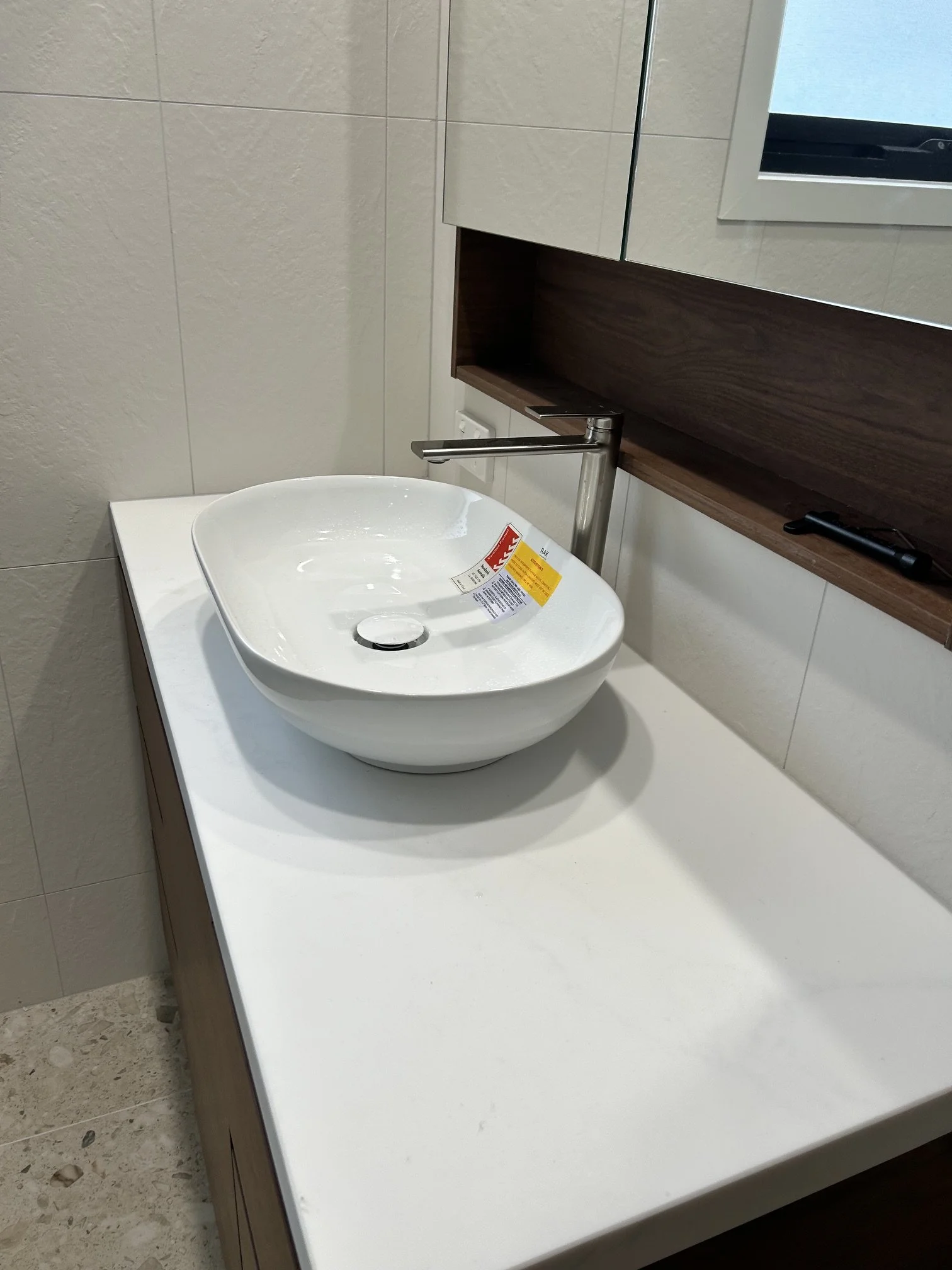 A bathroom vanity with a white ceramic vessel sink and a chrome faucet, mounted on a white countertop with a dark wood cabinet underneath, and a mirror and a window on the wall above.