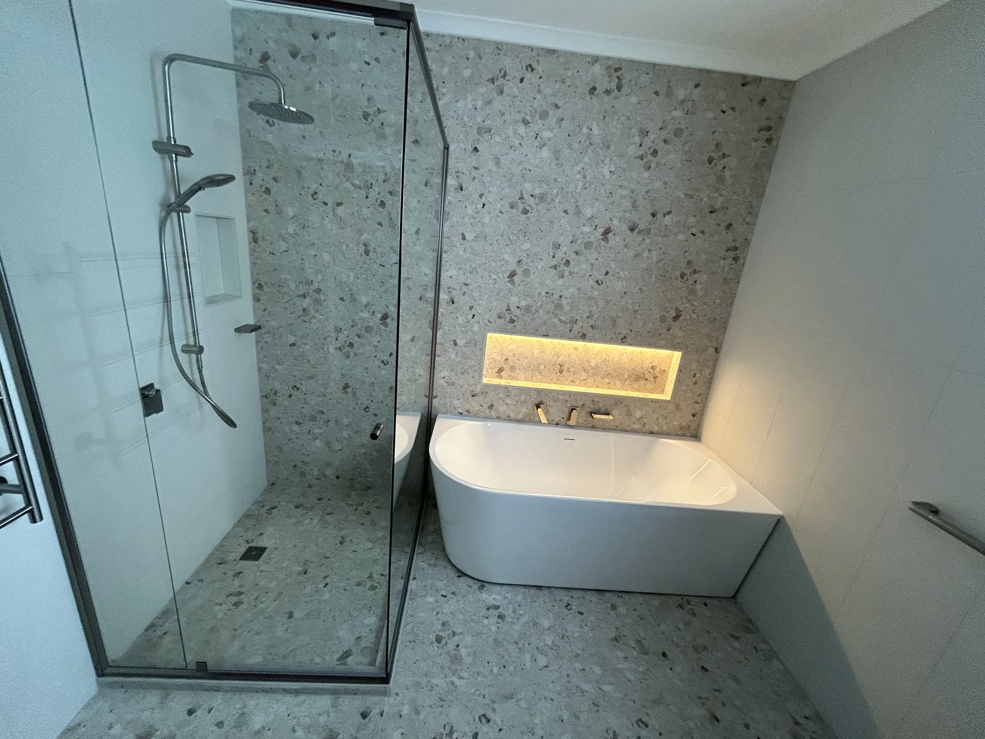 Modern bathroom with glass-enclosed shower, terrazzo wall and floor, and freestanding bathtub. There's a recessed lighting detail in the wall.