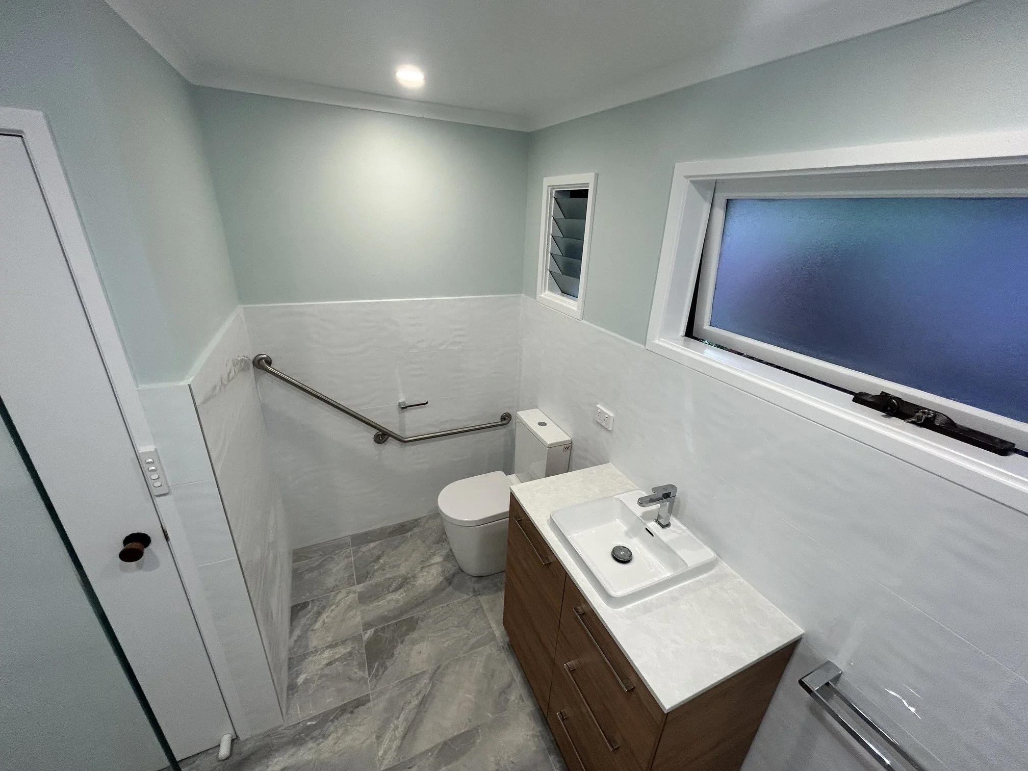 Modern accessible bathroom with wall-mounted grab rails, toilet, vanity sink, tiled walls, and frosted windows