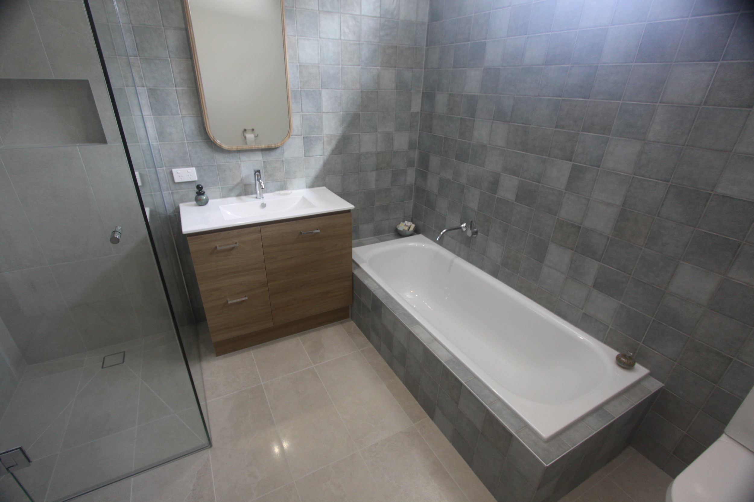 Modern gray-tiled bathroom with a white bathtub, glass shower enclosure, wood vanity, sink, and wall mirror