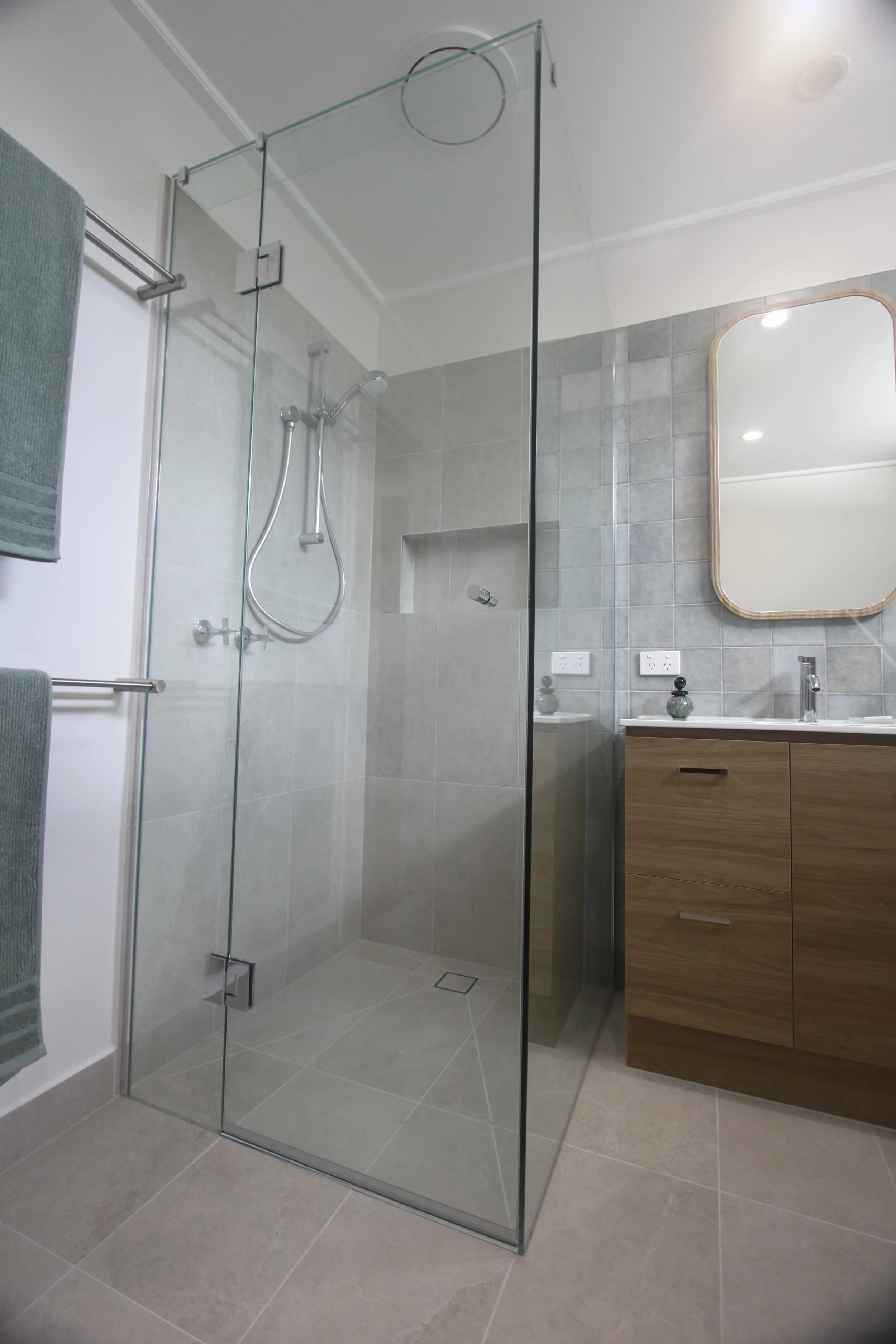 Modern bathroom with a frameless glass shower enclosure, gray tiled walls, wood vanity, wall mirror, and chrome fixtures.