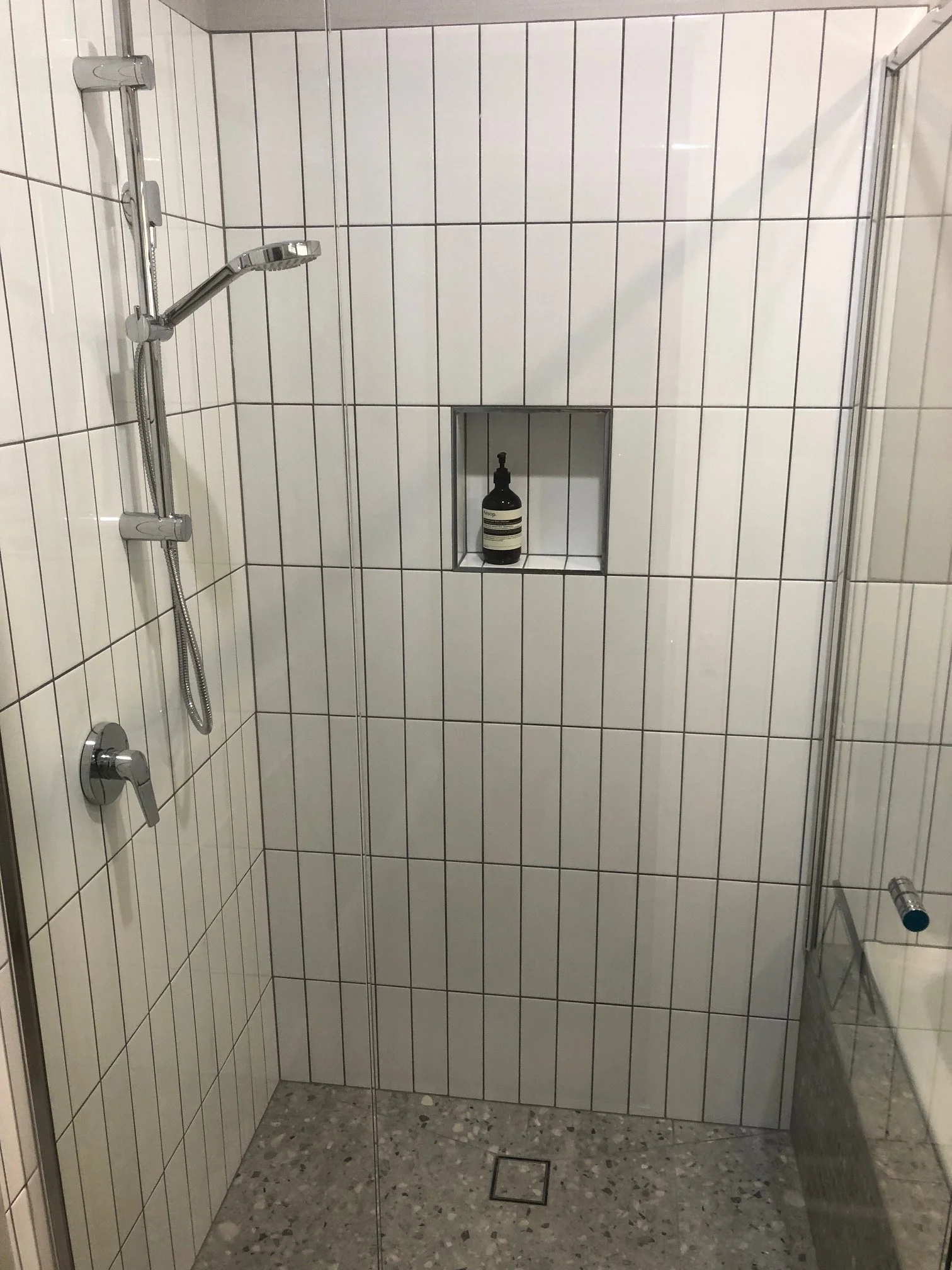Walk-in shower with white tiled walls, chrome handheld showerhead, glass door, recessed shelf.