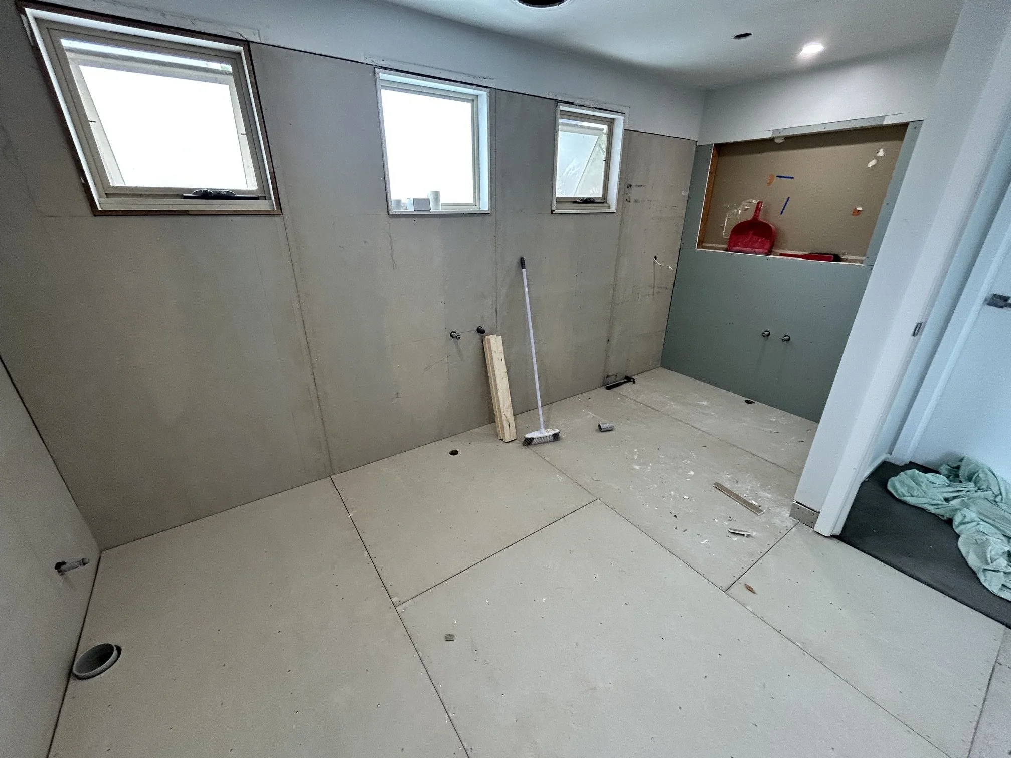 Unfinished bathroom under renovation with three windows, exposed plumbing, drywall, floor drains.