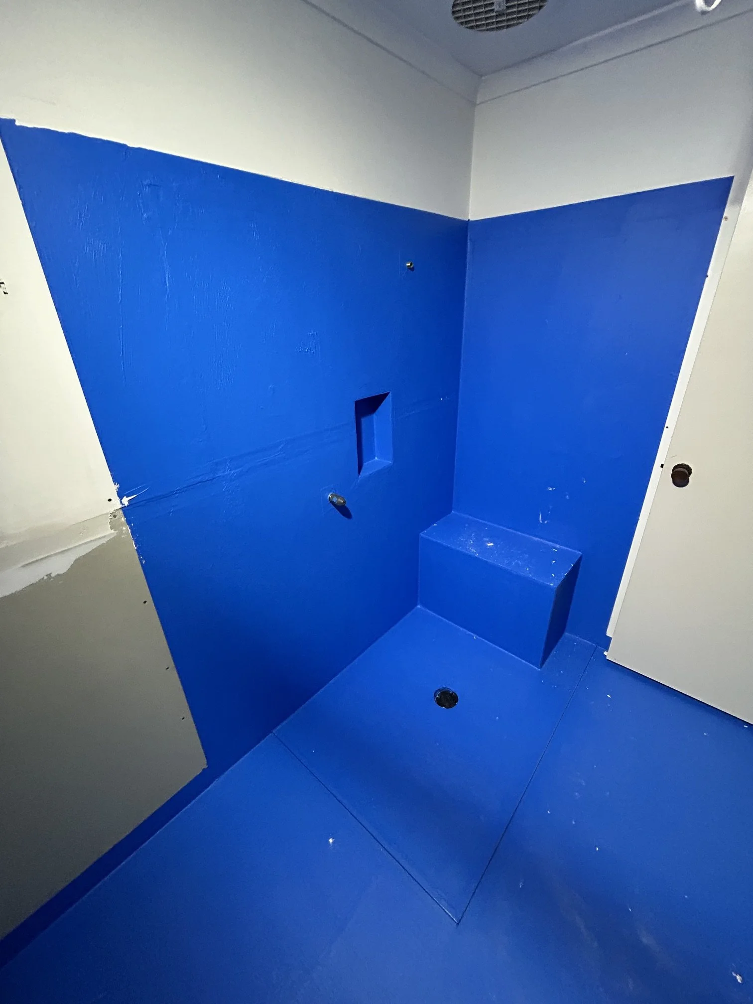Bright blue-painted bathroom stall with a raised step, wall fixtures, floor drain.