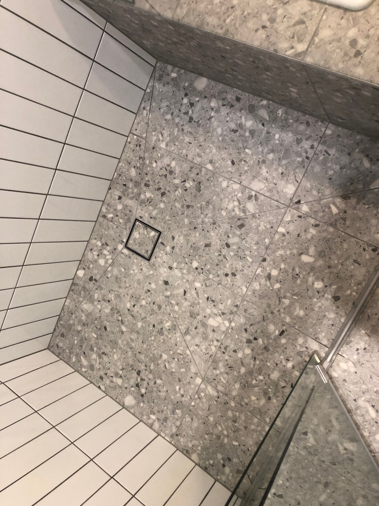 Speckled terrazzo tile floor beside a white tiled wall and glass shower enclosure