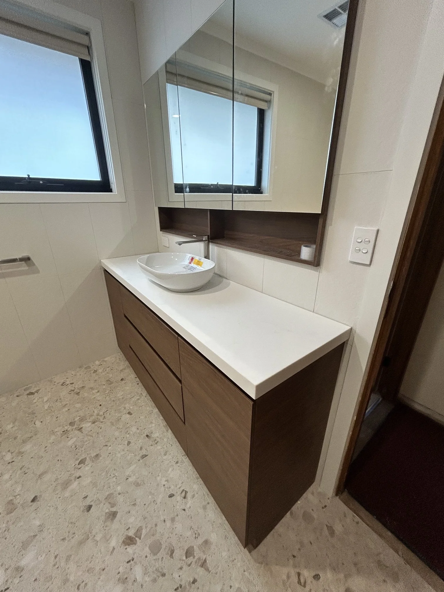 Modern bathroom with a white countertop, wooden cabinet drawers, a vessel sink, a large mirror, and a window with a white blind.