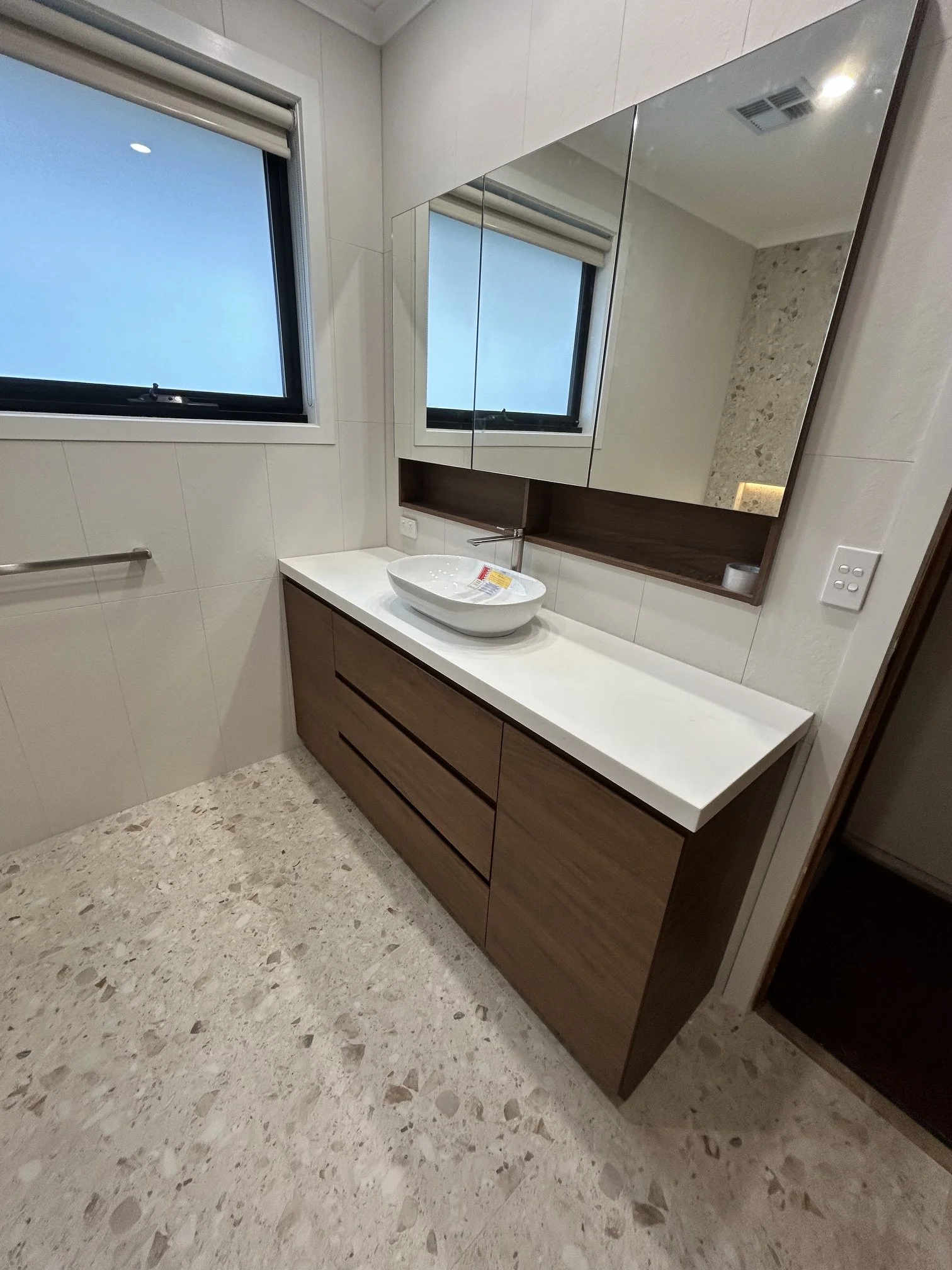 Modern bathroom interior with white walls, a window, a white countertop with a vessel sink, wooden cabinetry, a large mirror, and terrazzo-style flooring.