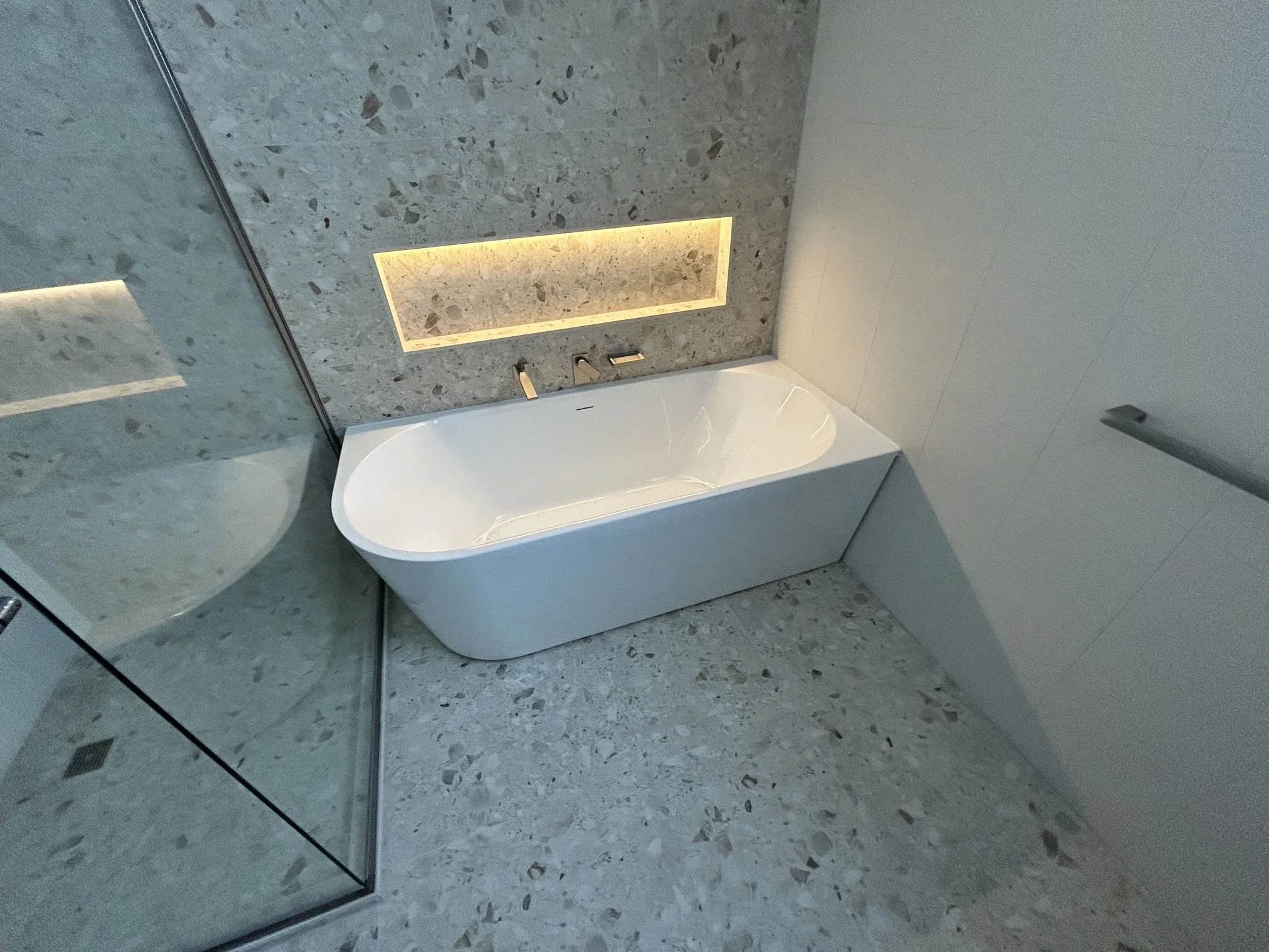Modern bathroom with terrazzo tile flooring, a white bathtub, and a glass shower enclosure with a metal frame. A rectangular niche with warm lighting is built into the tiled wall.