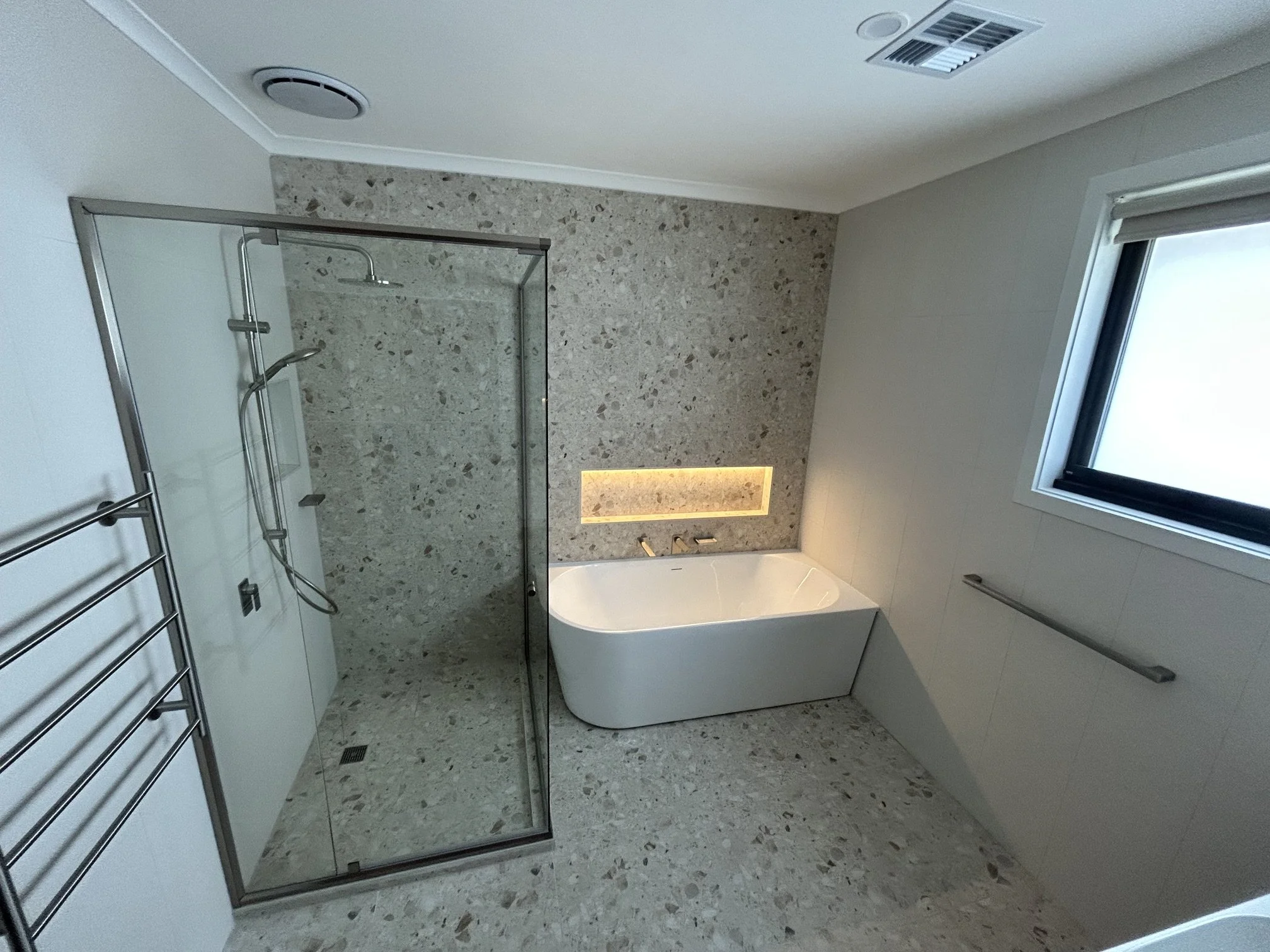 Modern bathroom with a glass shower enclosure, bathtub, window, and a tiled wall with a recessed shelf with LED lighting.