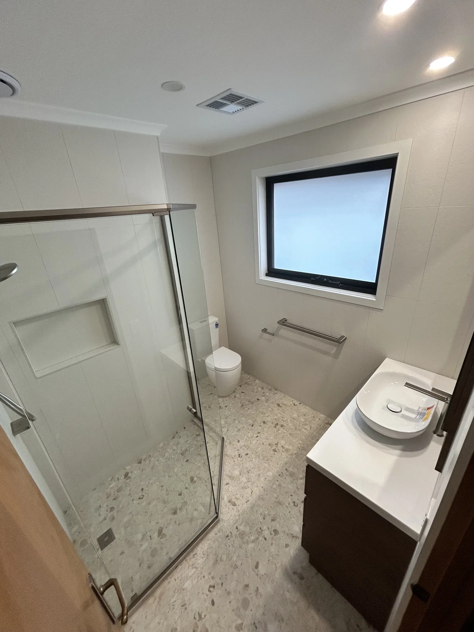 A modern bathroom with a glass shower enclosure on the left, a toilet in the corner, a small sink with a vessel basin on the right, a window above the toilet, beige tiled flooring, and recessed ceiling lighting.