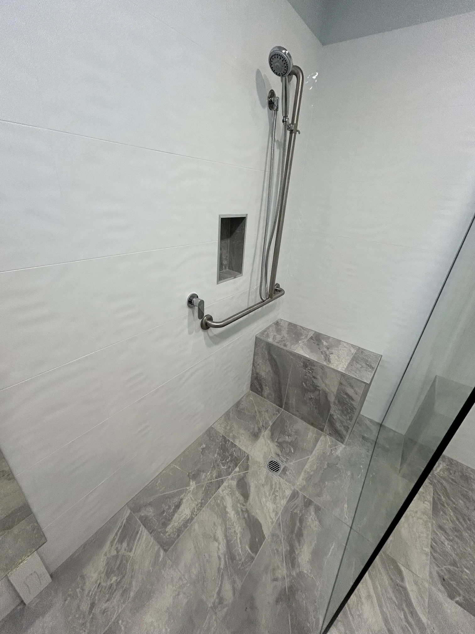 Modern walk-in shower with handheld showerhead, grab bar, built-in niche, bench seat, gray tile, and glass enclosure