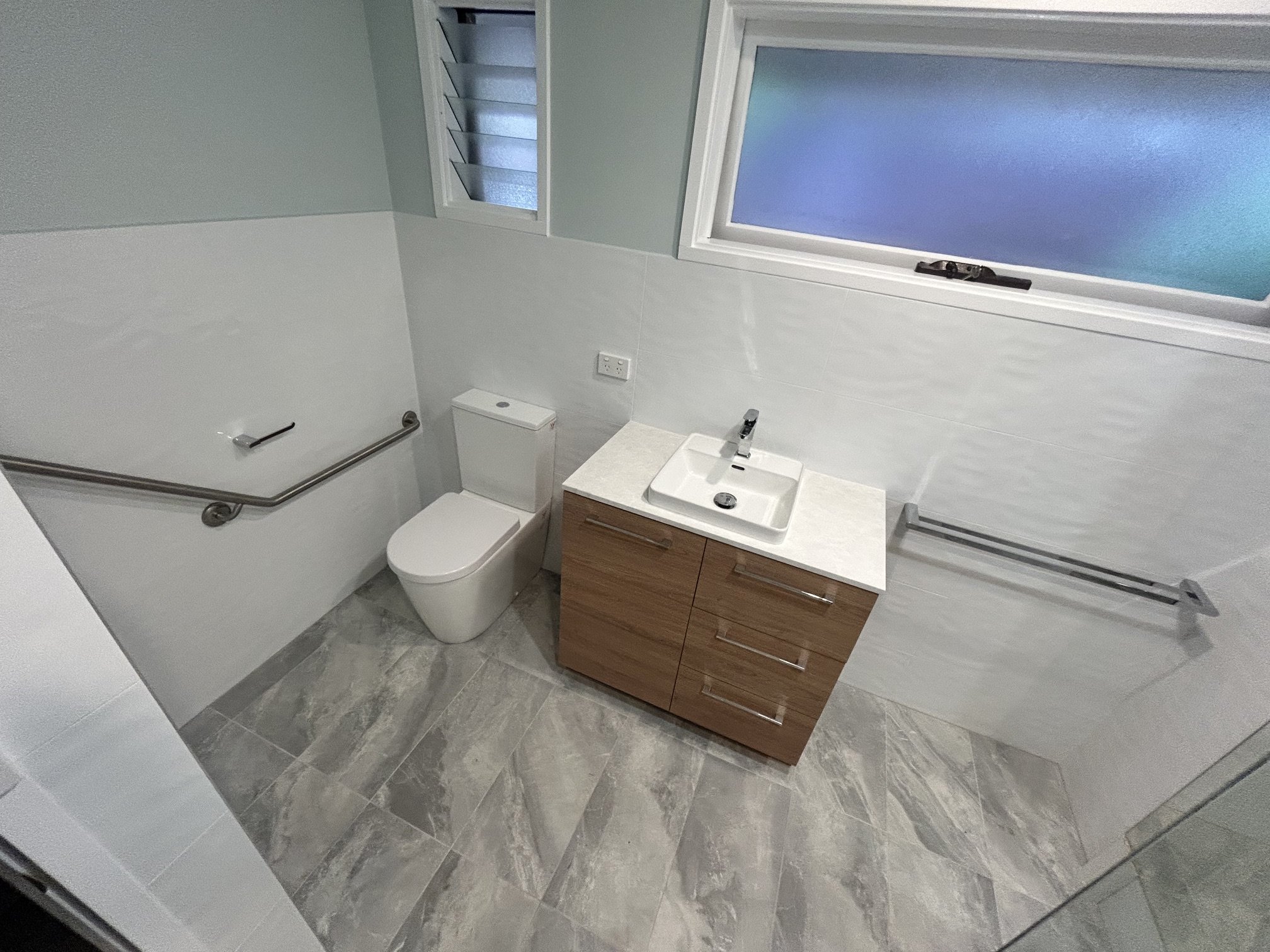 Modern white-tiled bathroom with toilet, wood vanity, rectangular sink, chrome grab rails, and frosted windows.