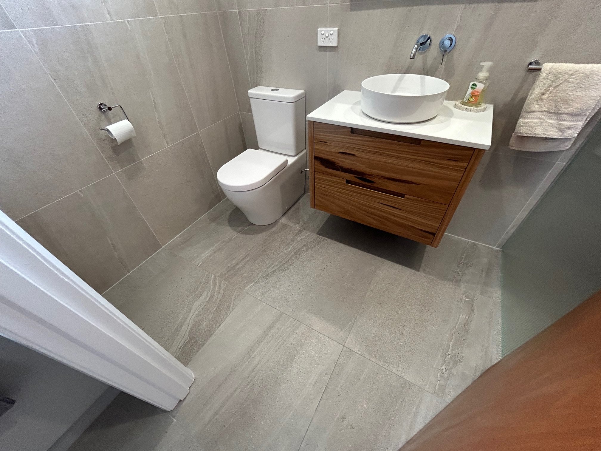 Modern tiled bathroom with a white toilet, wood vanity, vessel sink, wall-mounted faucets, towel, and toilet paper holder