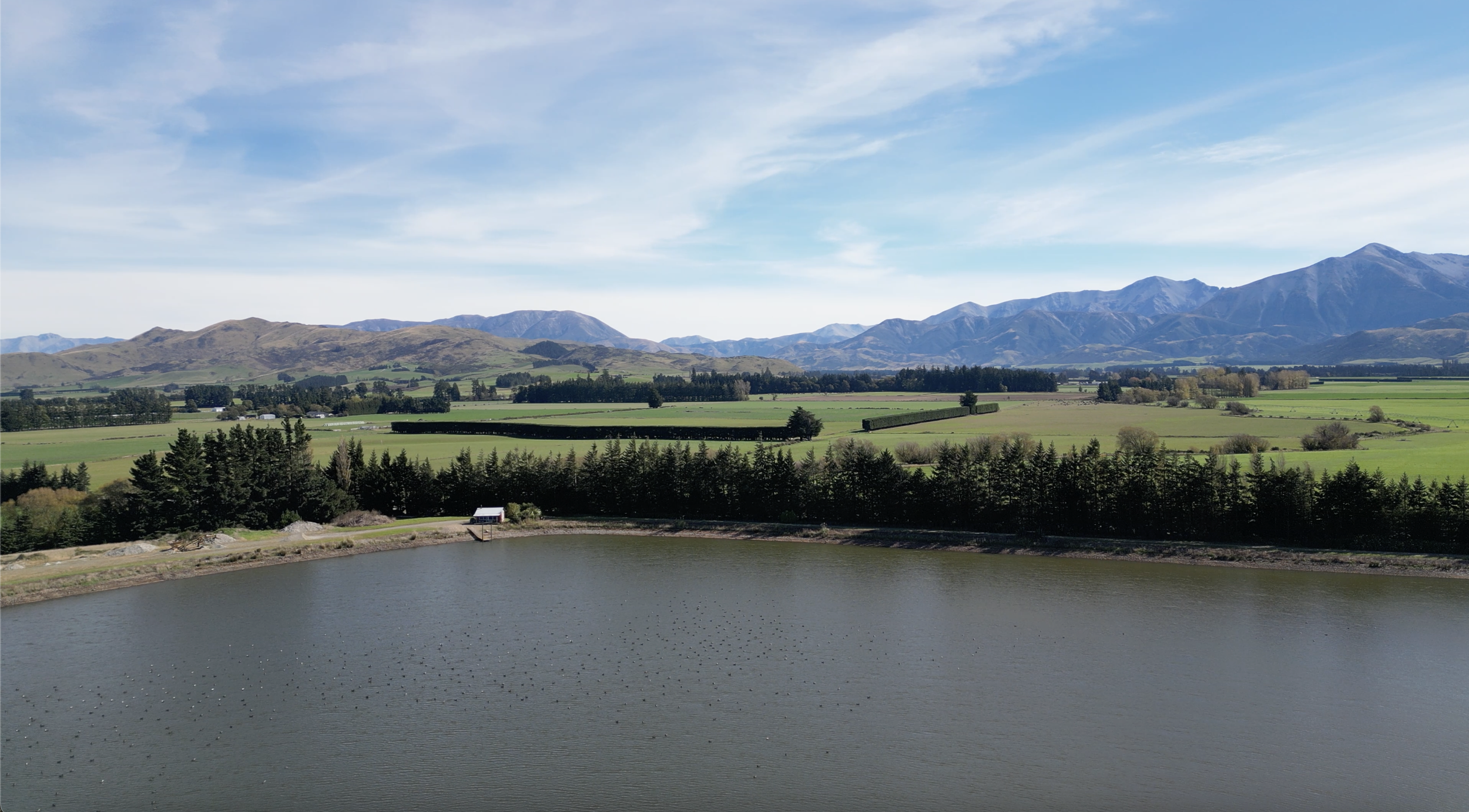 Scenic view of mountainous landscape with green fields, trees, a lake filled with grass carp, and a partly cloudy blue sky.