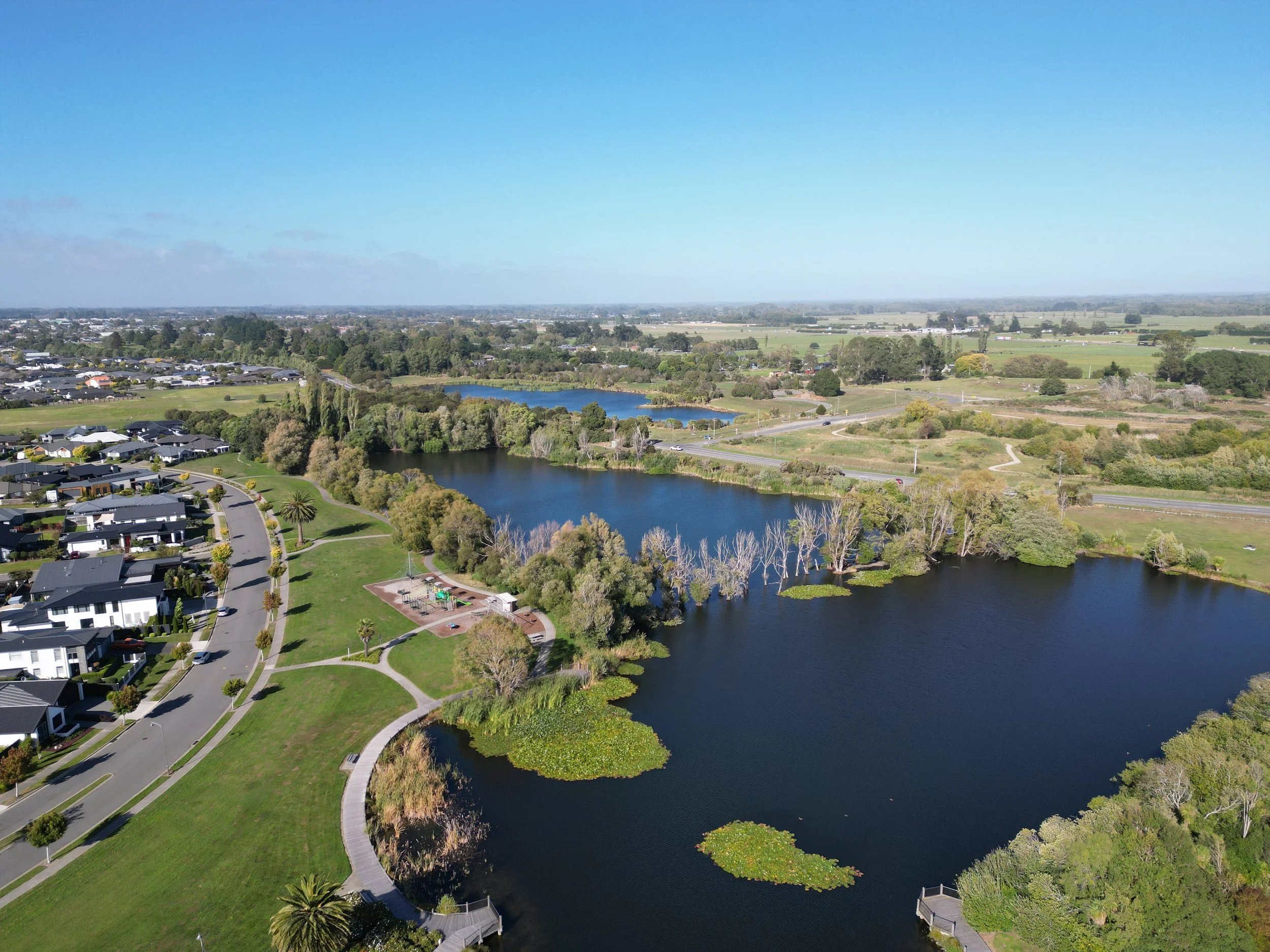 Aerial view of a suburban area with houses, trees, and a winding council owned lake in Kaiapoi, New Zealand under a clear blue sky.