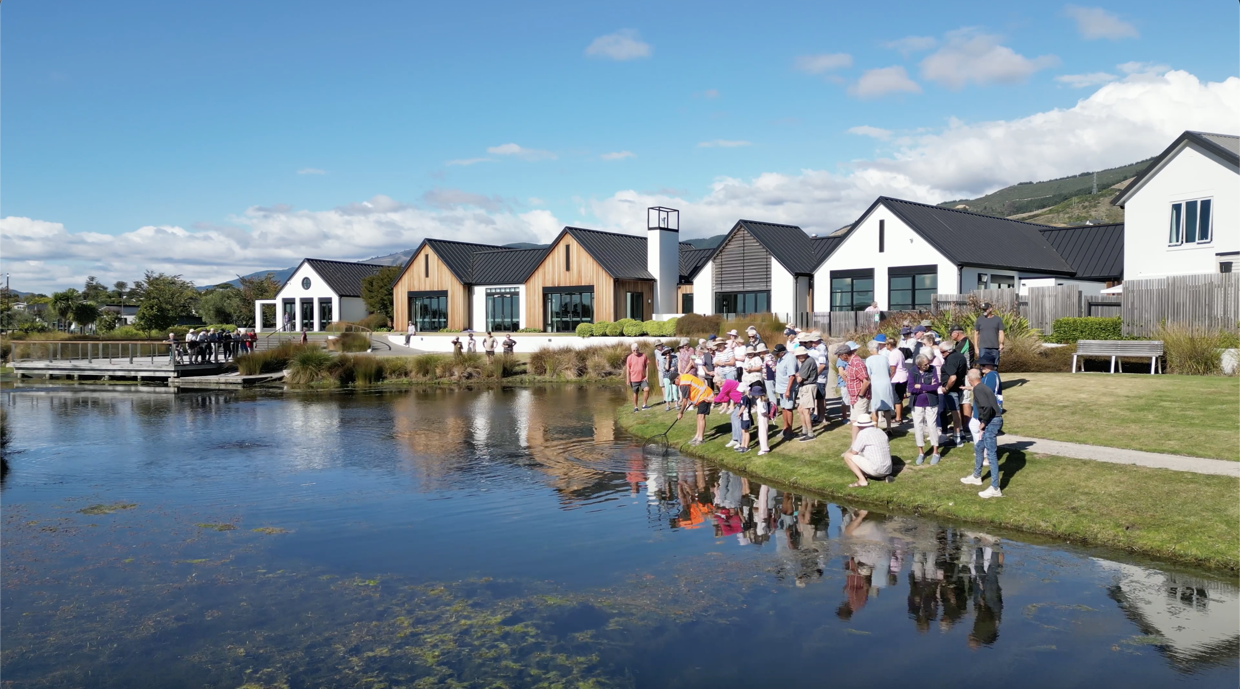 People gather near a pond with reflections of buildings and trees, in front of retirement village rest home on a sunny day with a partly cloudy sky.