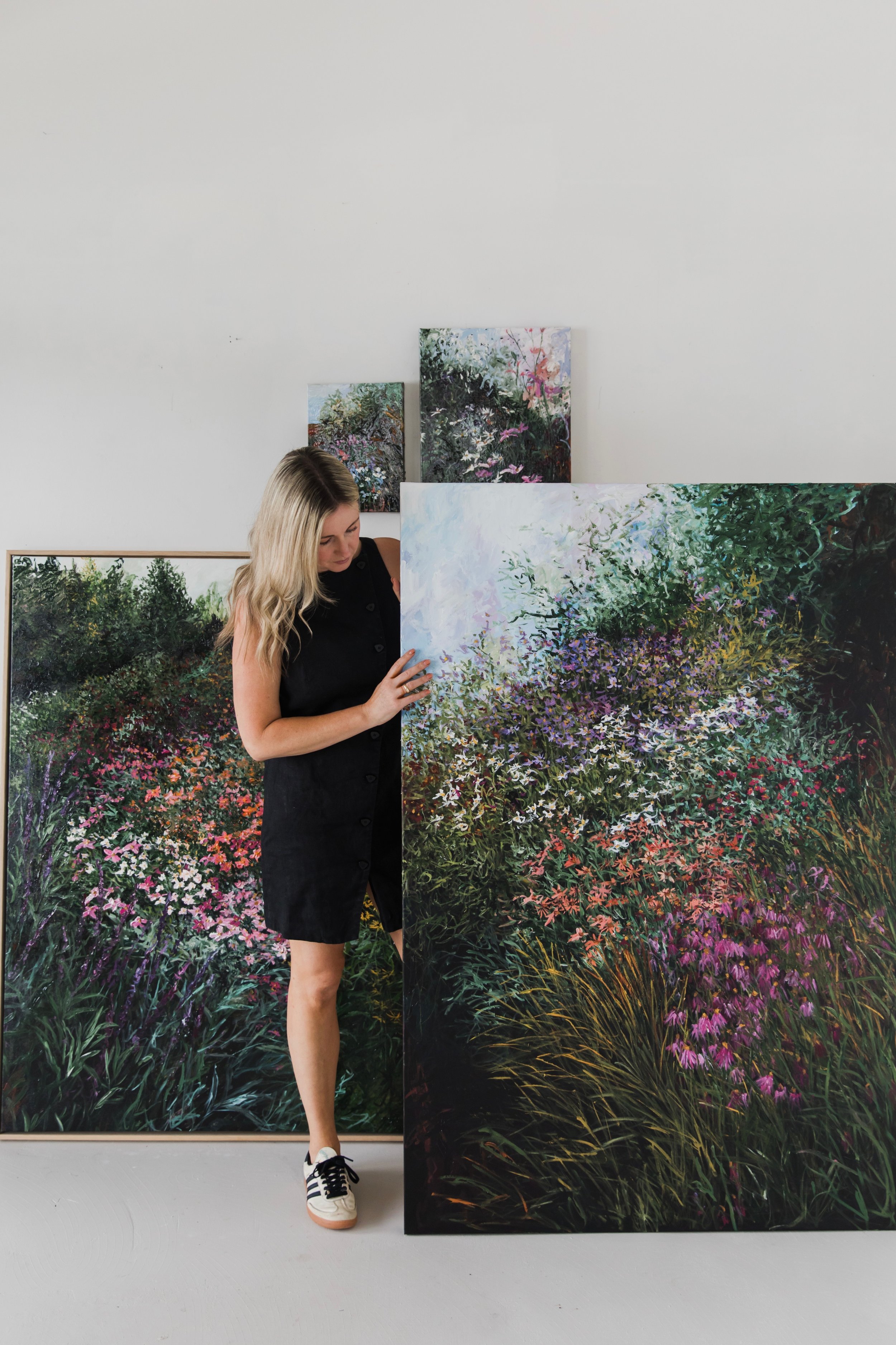 A woman with long blonde hair in a black sleeveless dress and white sneakers is standing in front of large colorful paintings of garden scenes, holding one of the paintings.