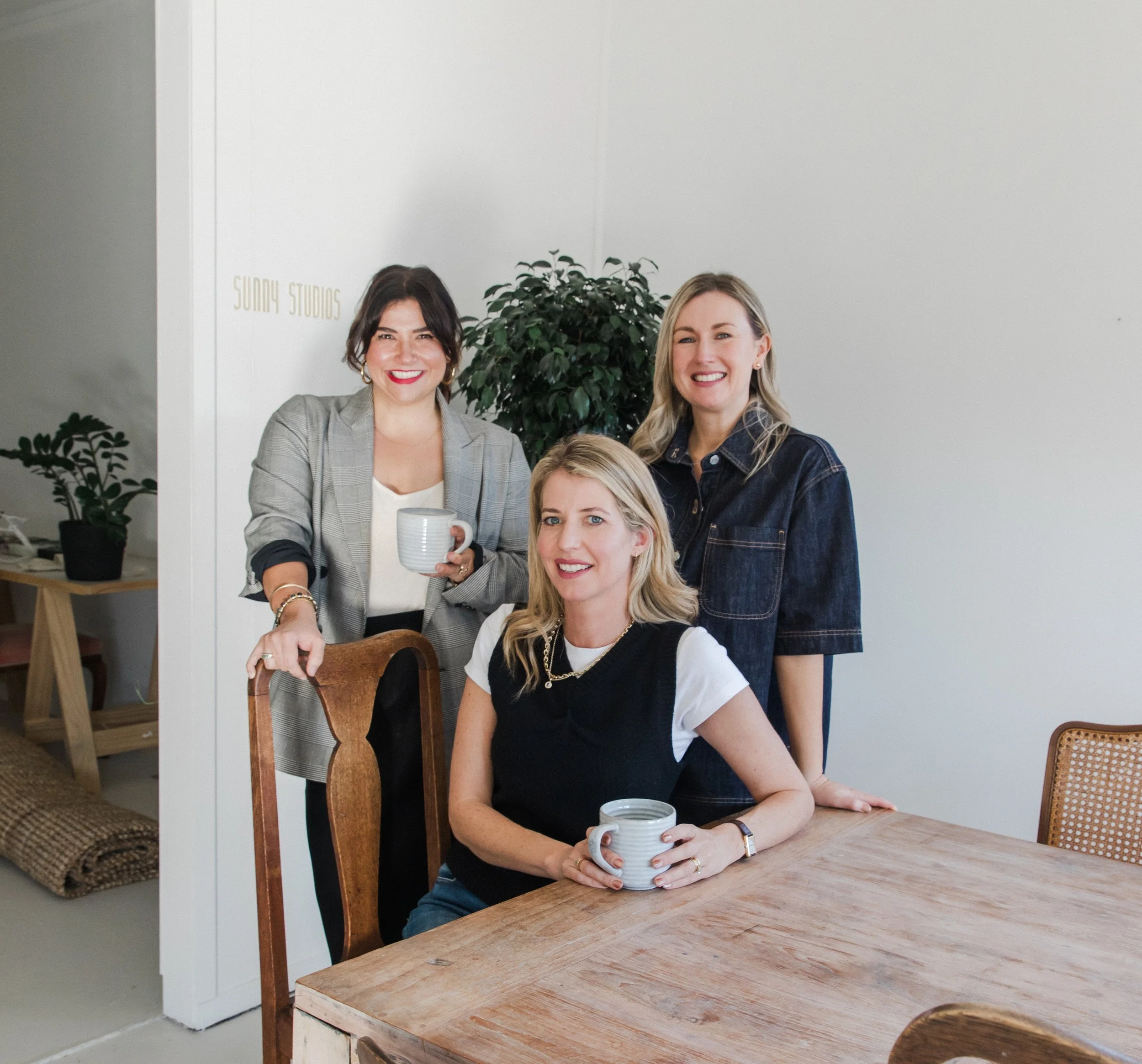 Four women smiling around a wooden table, all holding coffee mugs, with potted plants and a white wall in the background that has a sign reading 'Sunny Studios.'