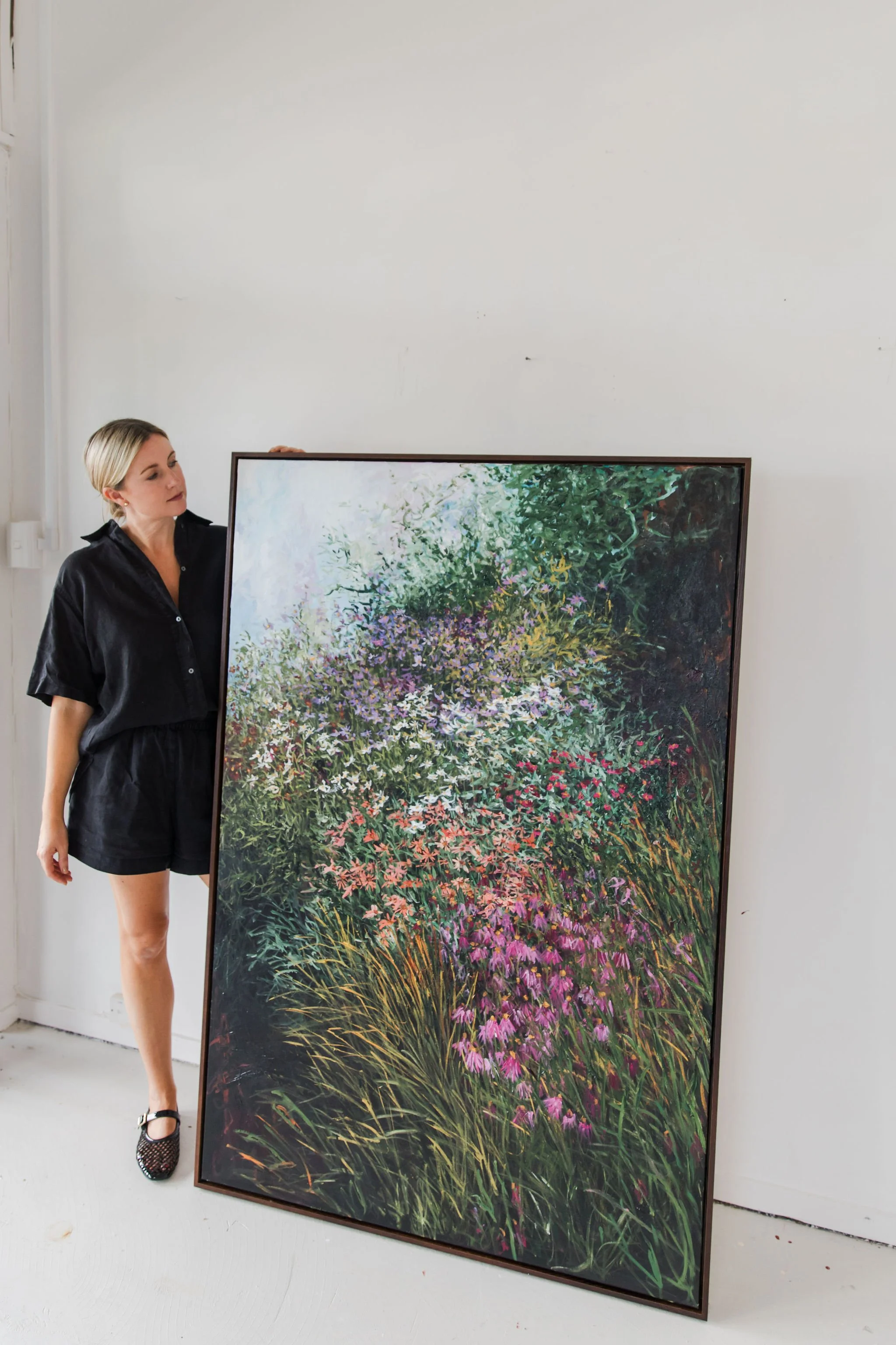 A woman with blonde hair wearing a black outfit and black shoes with a pattern, holding a large framed colorful floral painting, standing against a plain white wall.