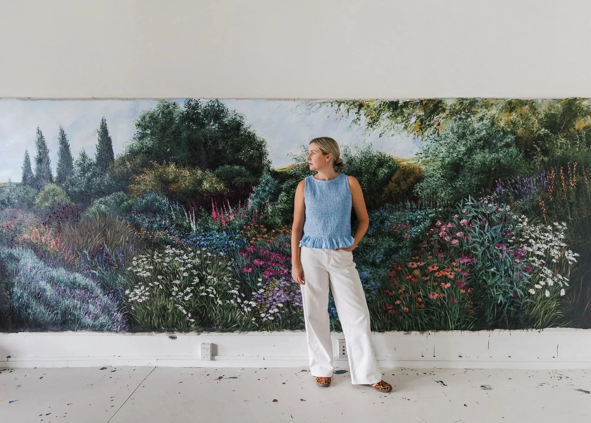 A woman standing in front of a large floral painting, wearing a light blue sleeveless top, white wide-leg pants, and animal print shoes, with her hands in her pockets.