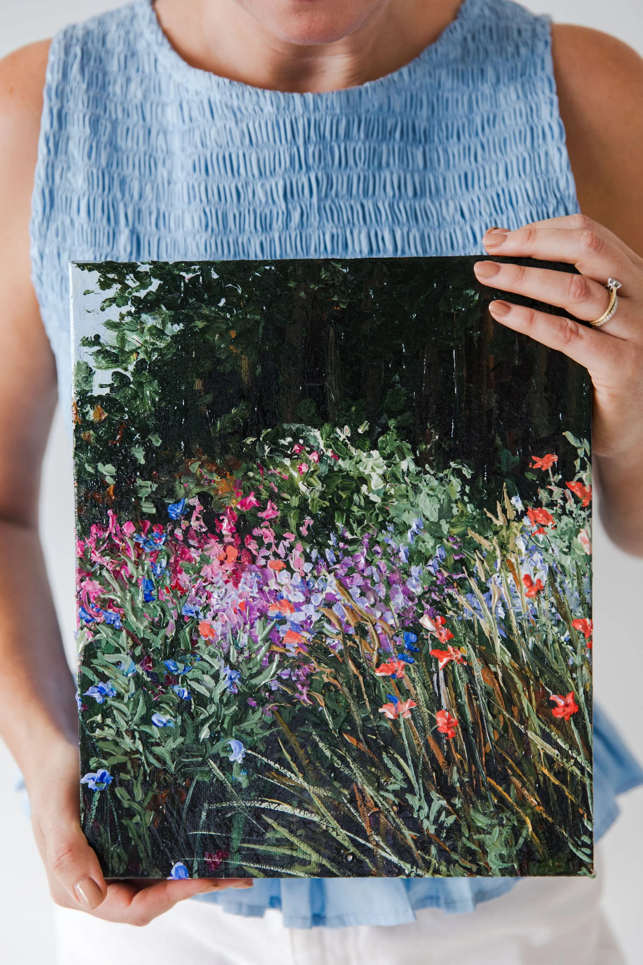 A person wearing a sleeveless light blue top holding a painting of a colorful garden scene with green foliage and vibrant flowers.