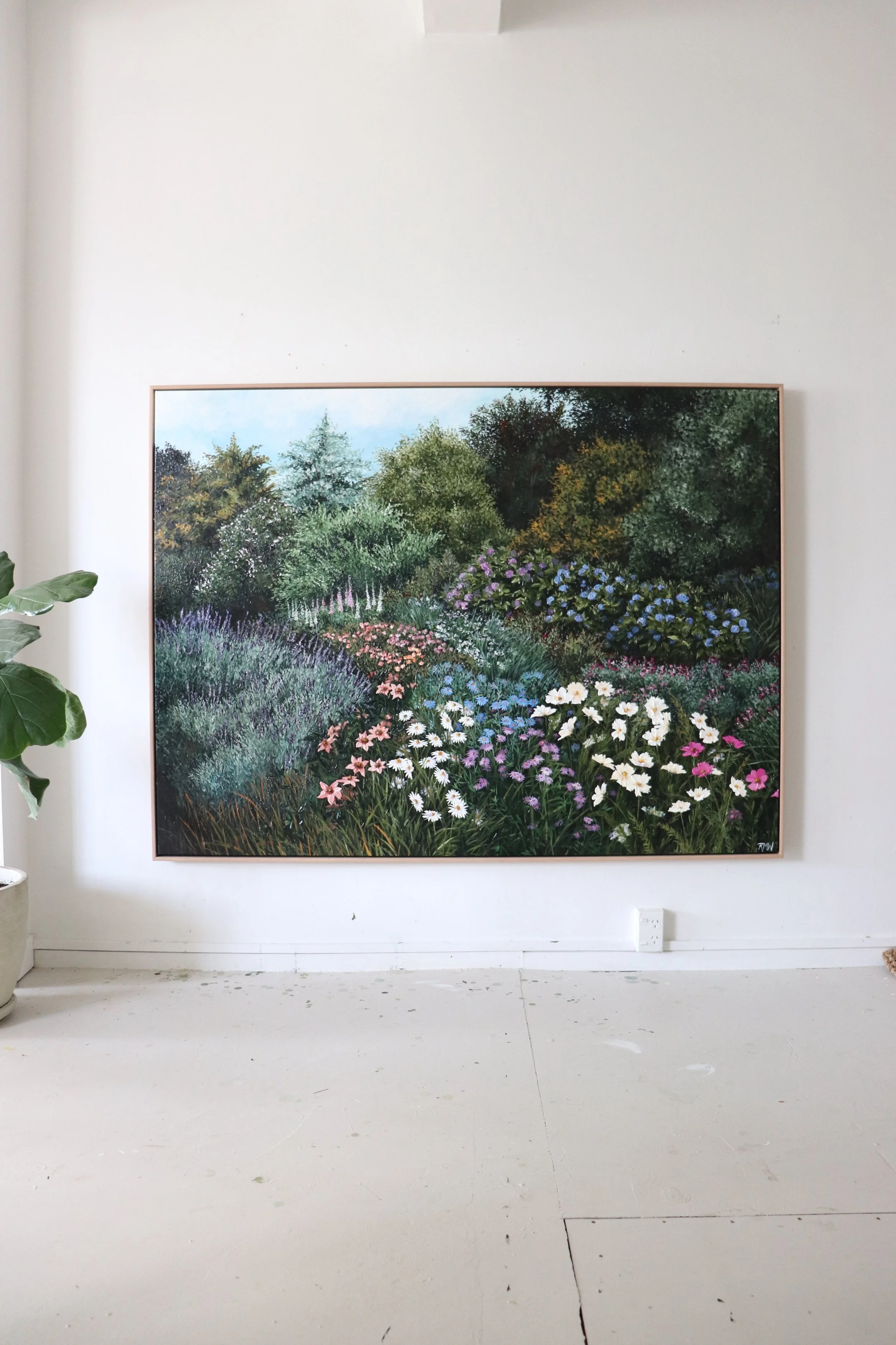 A large framed garden landscape painting hanging on a white wall in a gallery. The painting depicts a lush garden with a variety of colorful flowers and dense green trees.