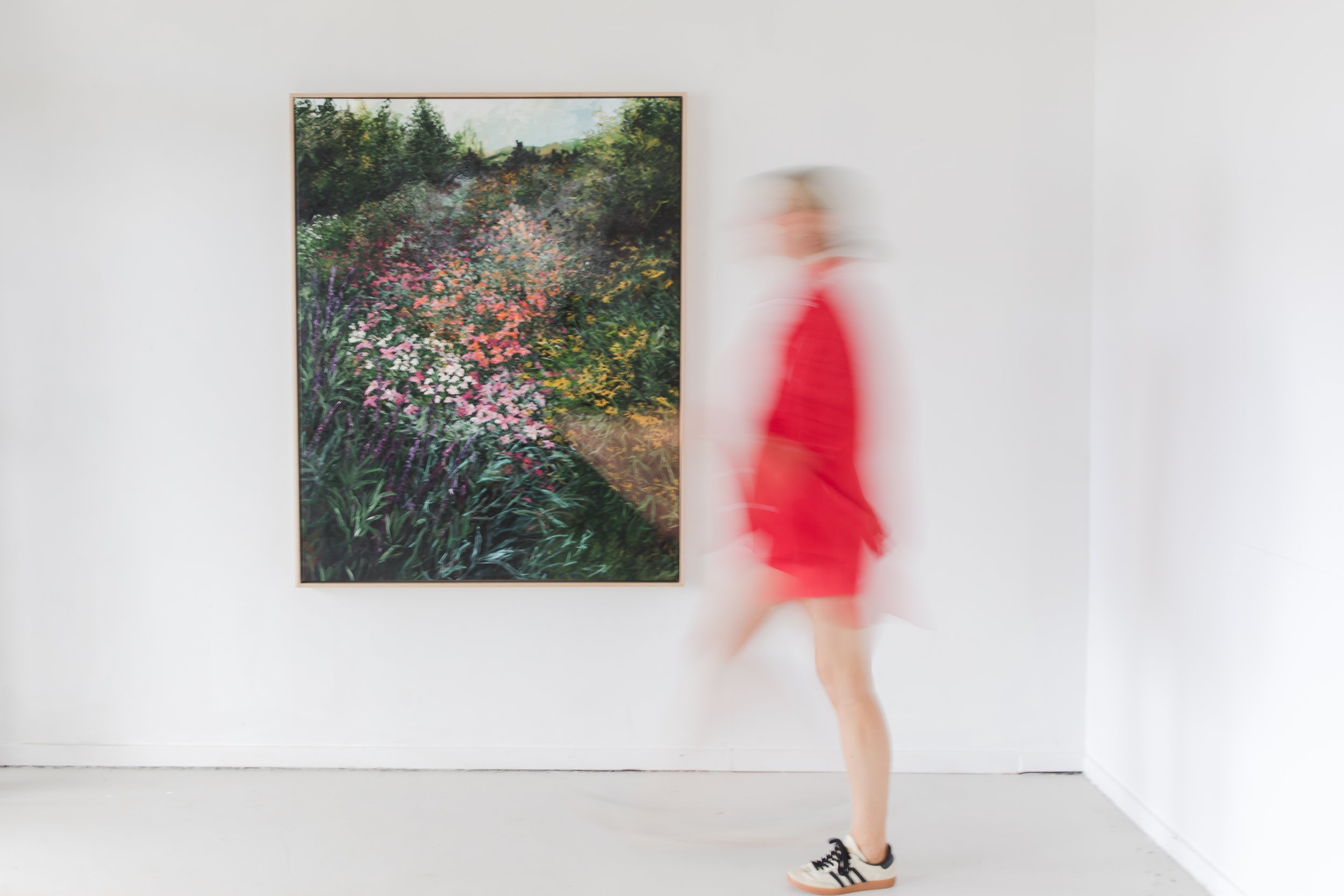 A person in a red dress and sneakers walking past a framed painting of a colorful garden on a white gallery wall.