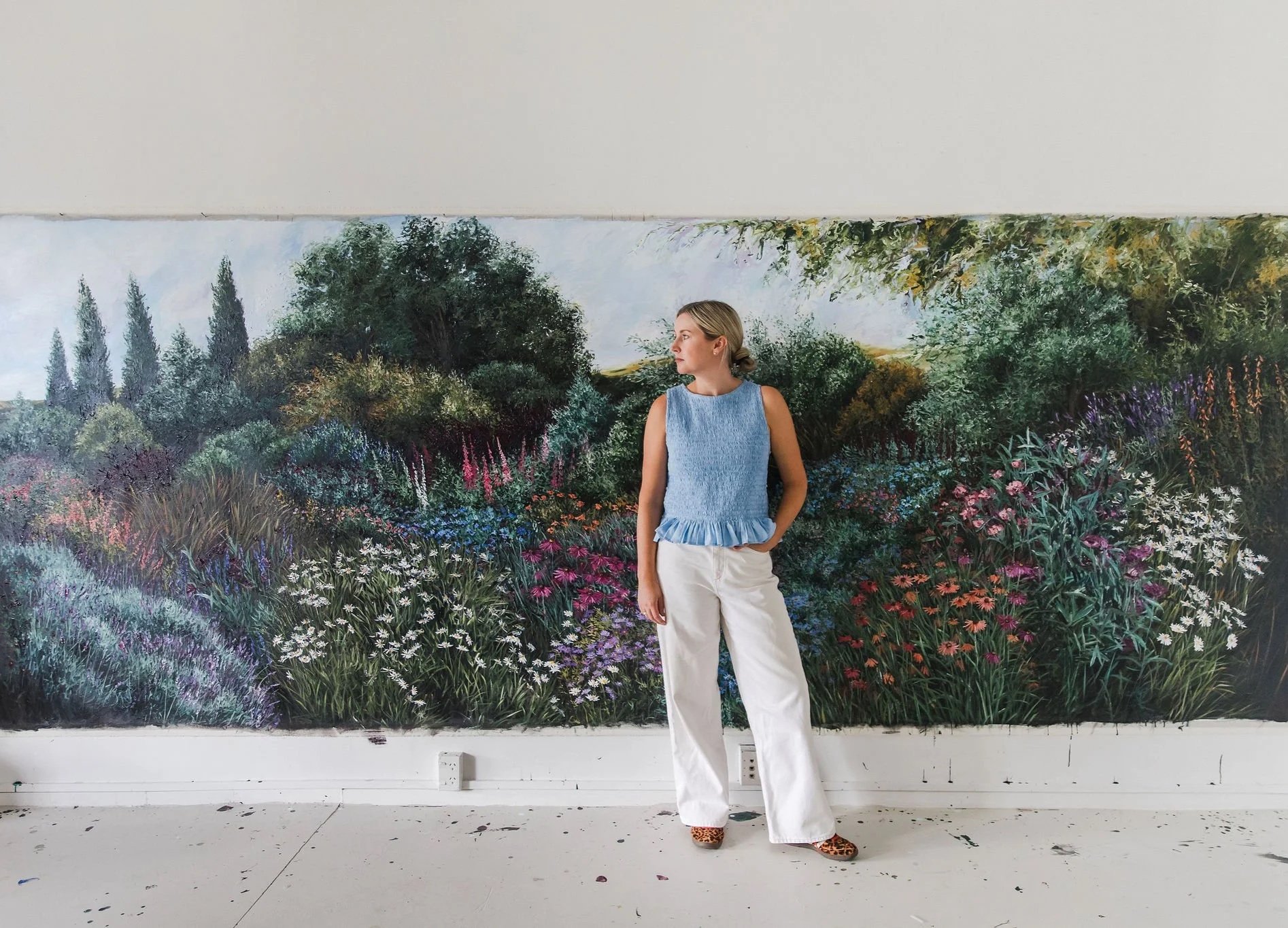 A woman stands in front of a large detailed painting of a lush garden with various trees and colorful flowers. She is wearing a blue sleeveless top, white pants, and leopard print shoes.