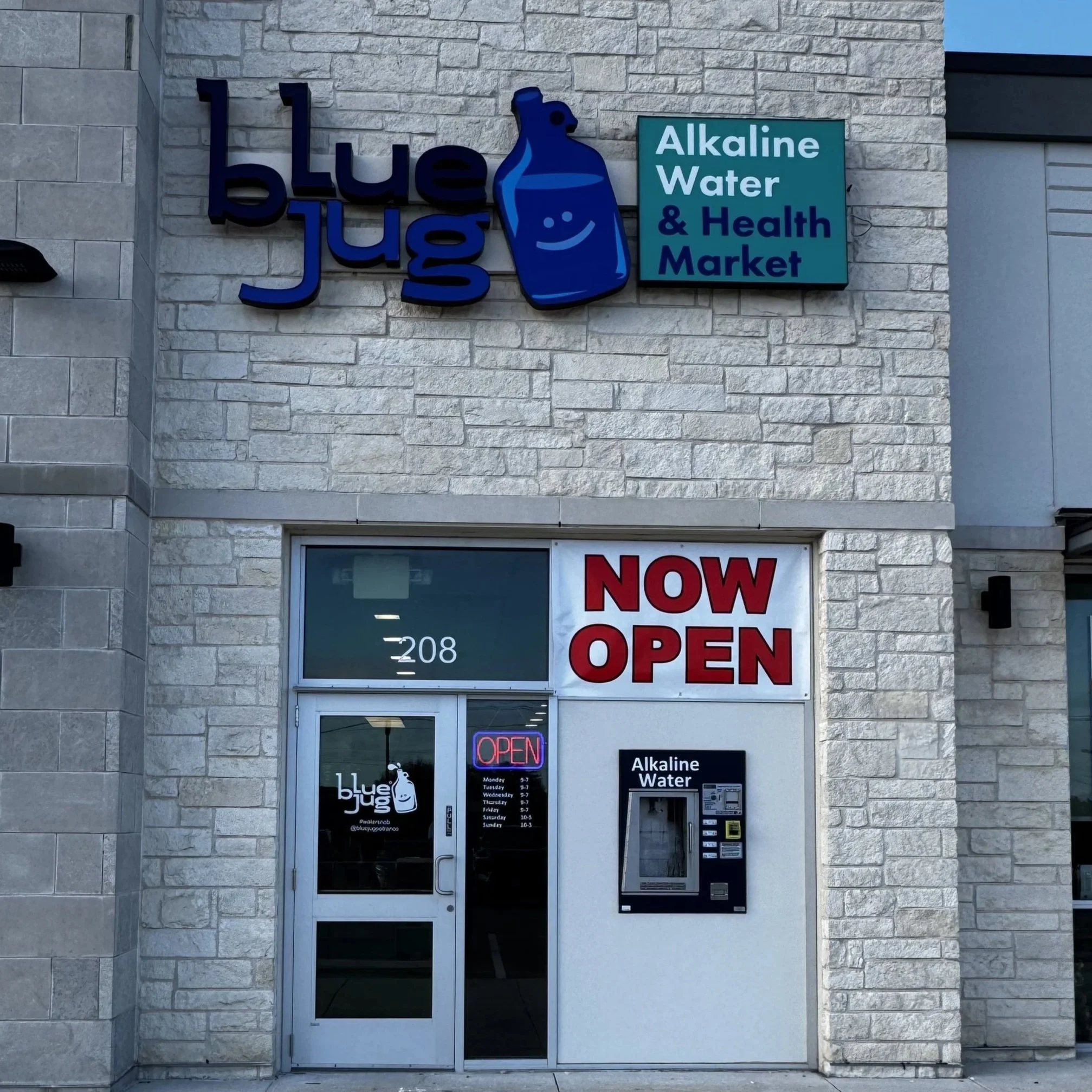 Storefront with signs for alkaline water and health market, and a digital and paper menu, with a 'Now Open' sign in the window.