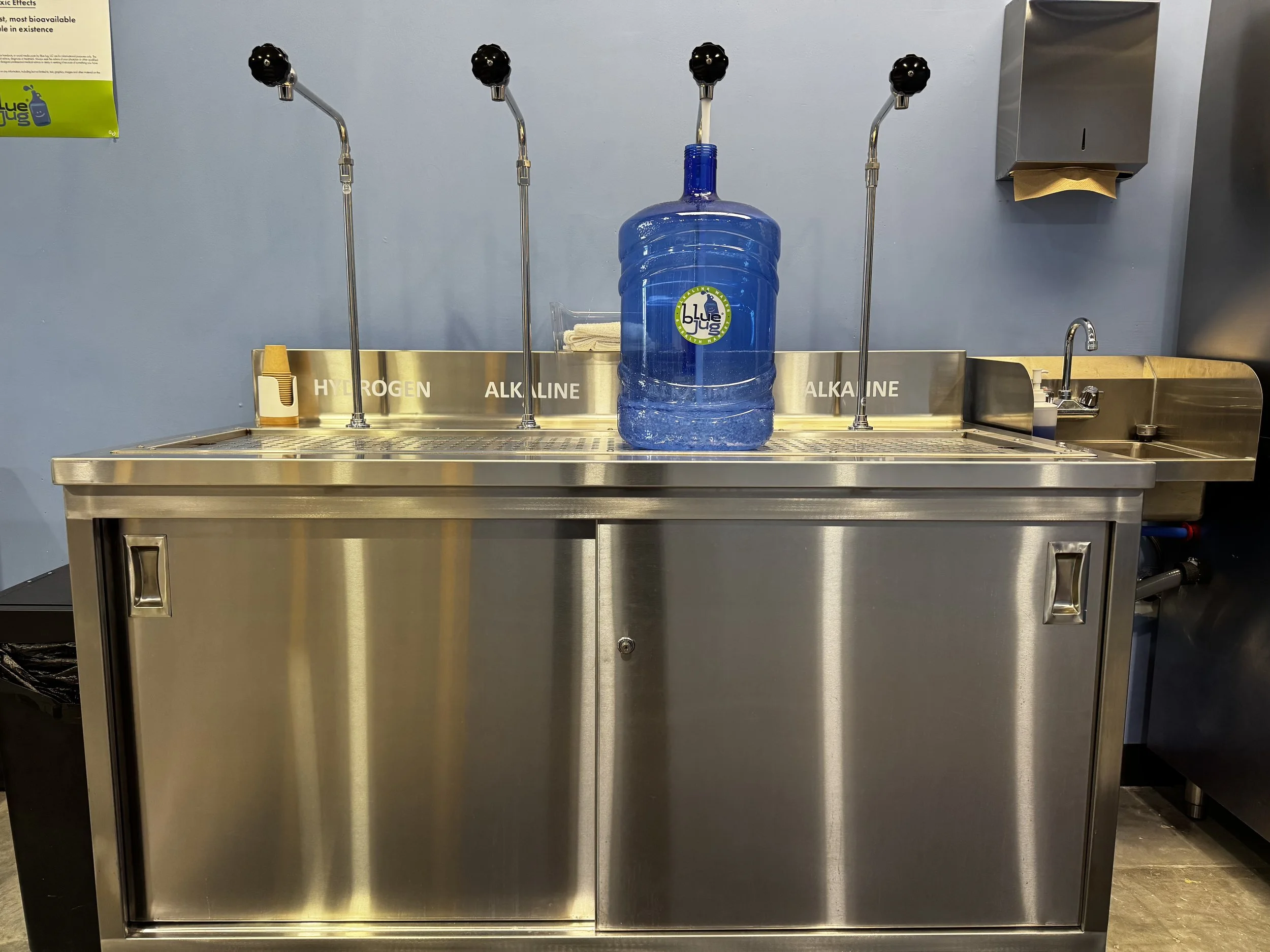 A stainless steel water station with four nozzles labeled for hydrogen, alkaline, and other water types. A large blue water jug is placed on top. To the right, there is a paper towel dispenser and a small sink with a faucet.