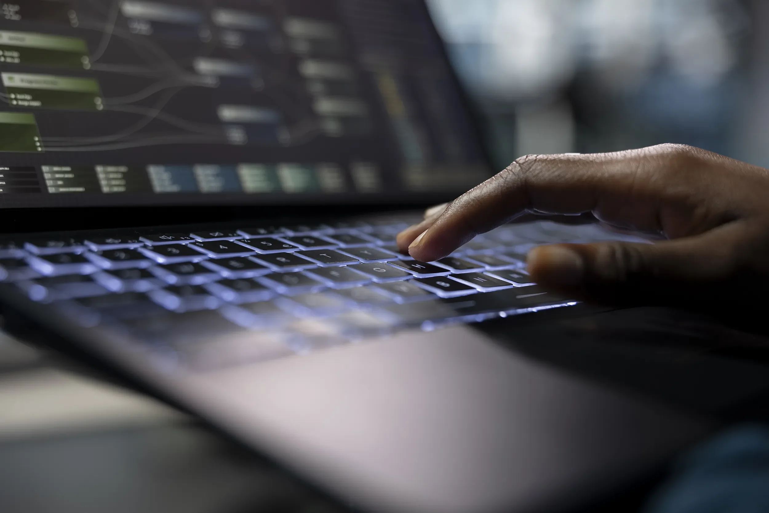 Close-up of a person's hand typing on a laptop keyboard with a blurred screen showing data or graphical interface.