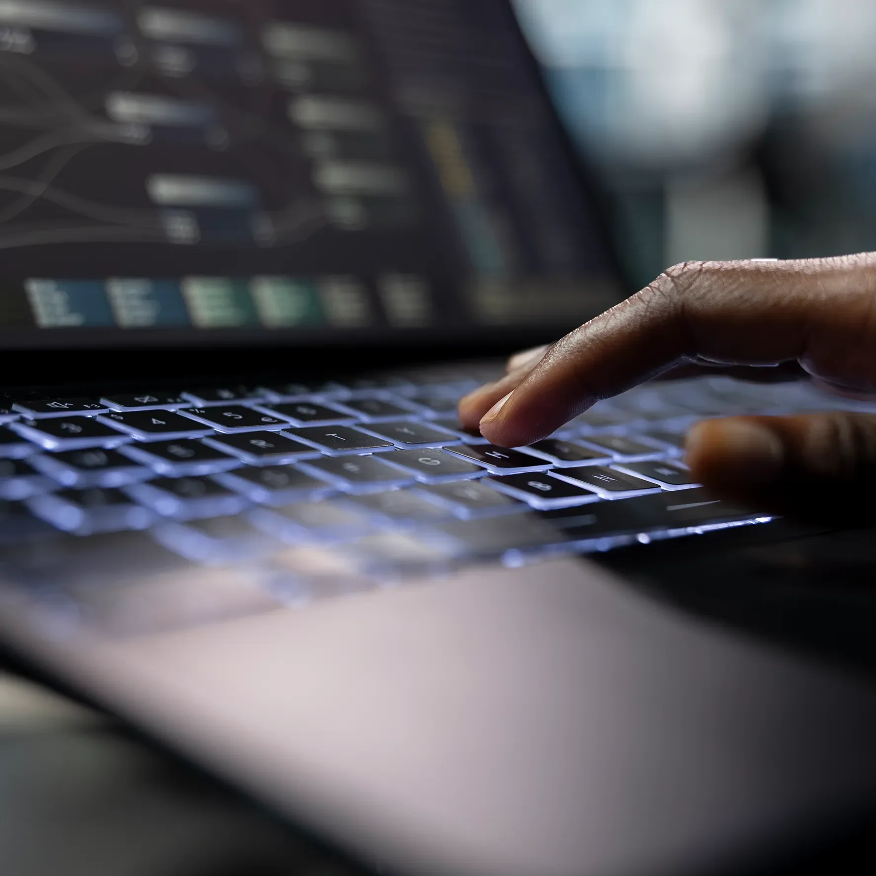 Close-up of a person's hand typing on a laptop keyboard with a blurry computer screen in the background.