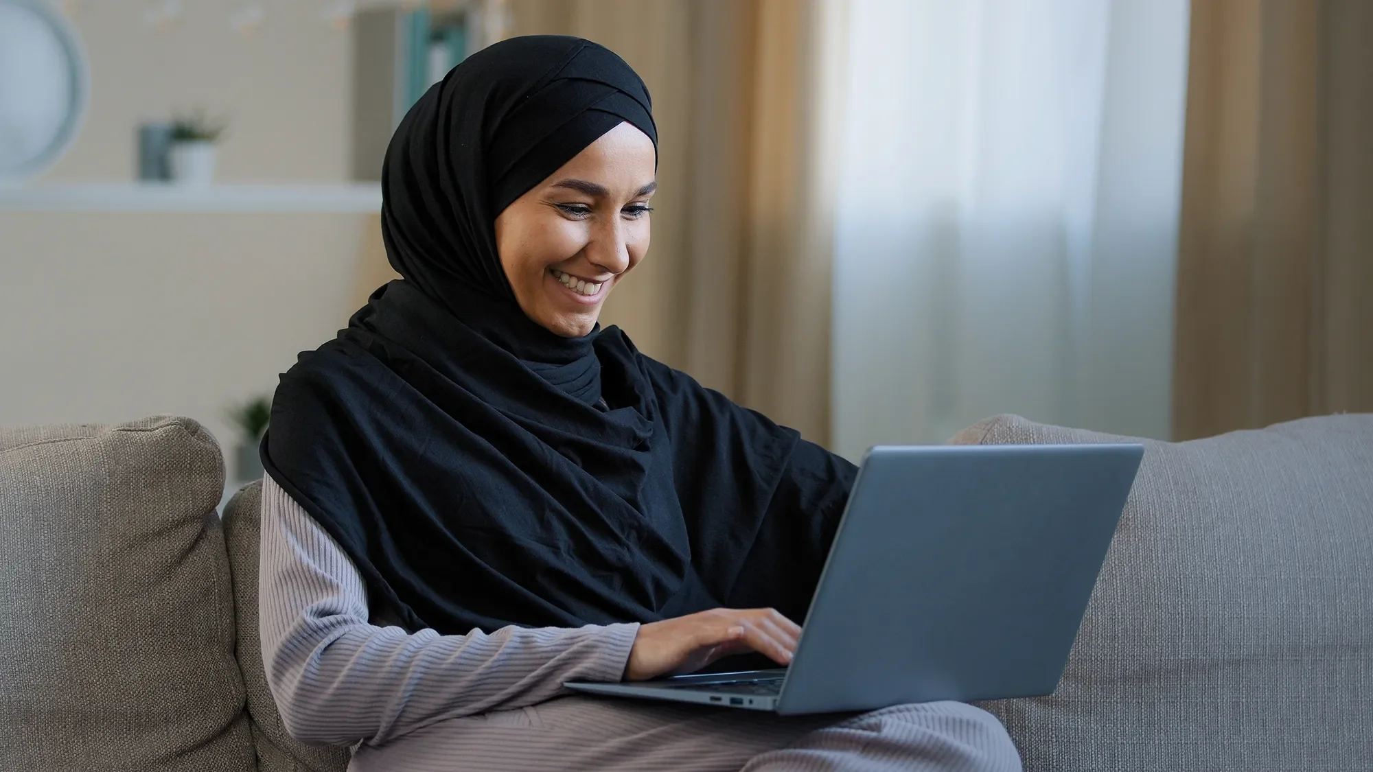 A young woman in a black hijab and striped long sleeve shirt sitting on a beige sofa using a silver laptop in a cozy living room with beige curtains and a small potted plant on a shelf in the background.