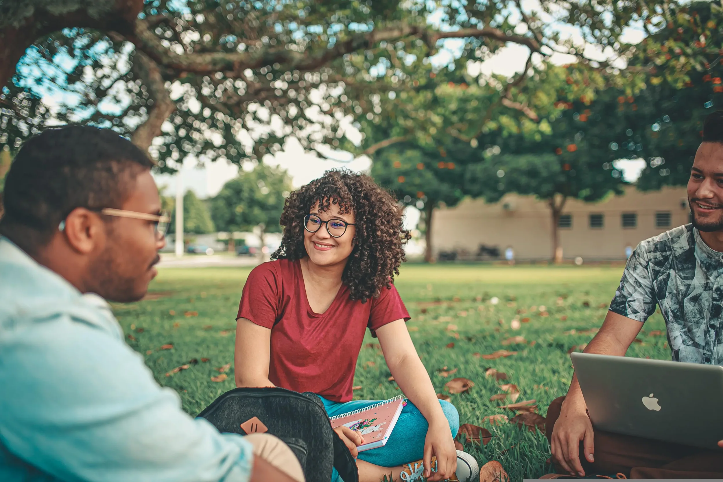 Four young adults sitting on grass in a park, engaging in conversation and smiling. There are trees and a building in the background, and they have various notebooks and a laptop with an apple logo.
