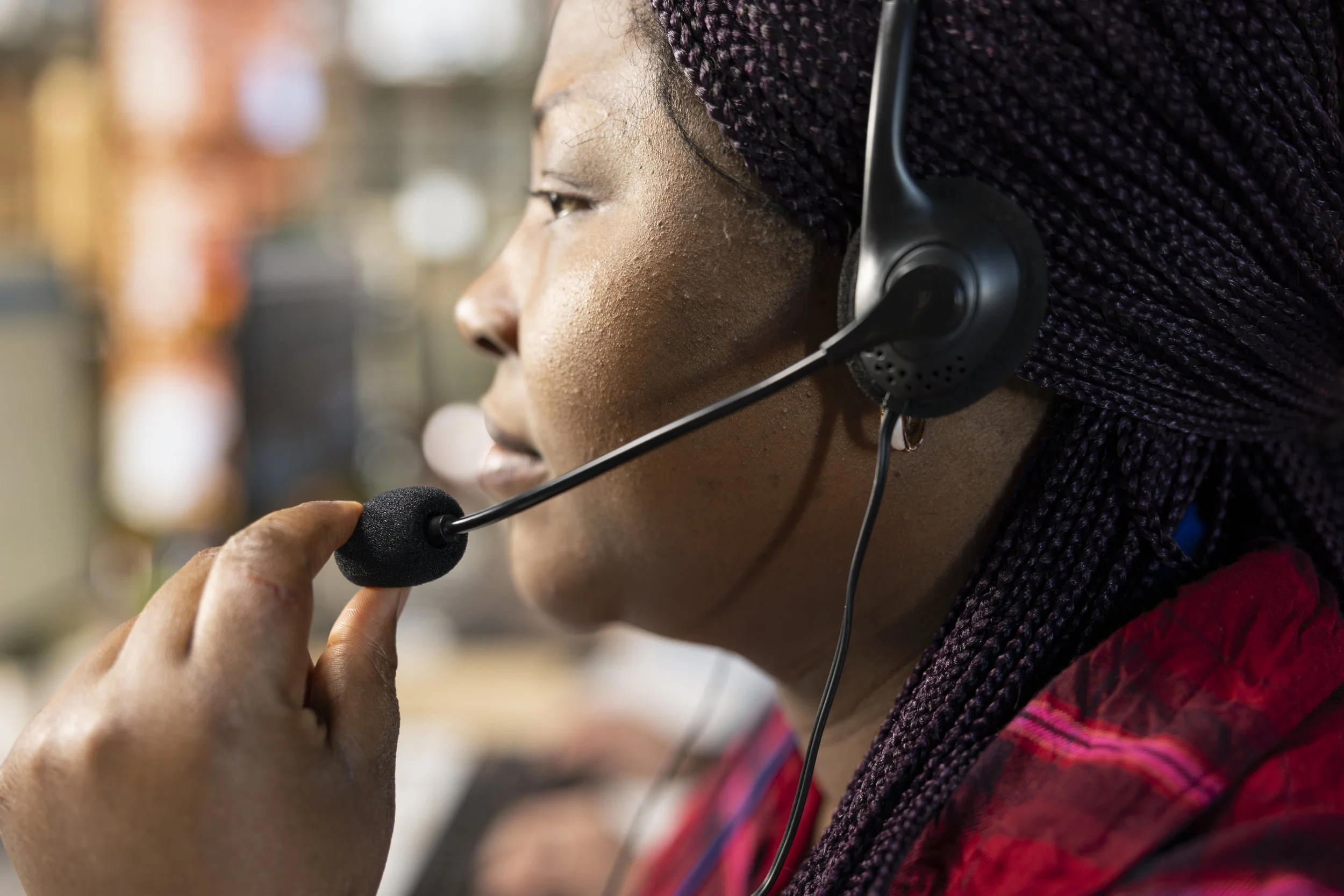 Close-up of a woman with braided hair wearing a headset microphone, speaking into the microphone in an indoor setting.