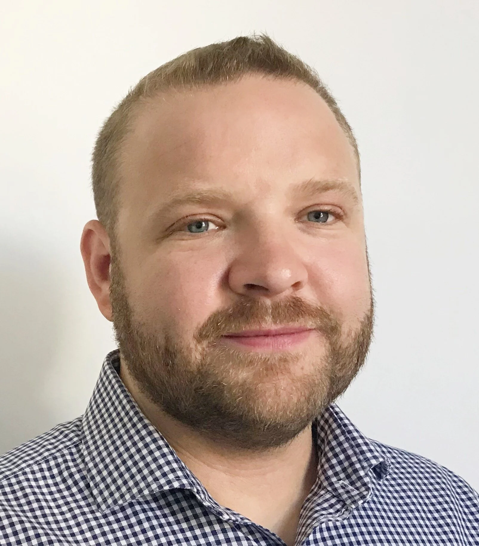 Close-up of a smiling man with blue eyes, light brown hair, and a beard, wearing a checkered shirt against a plain white background.